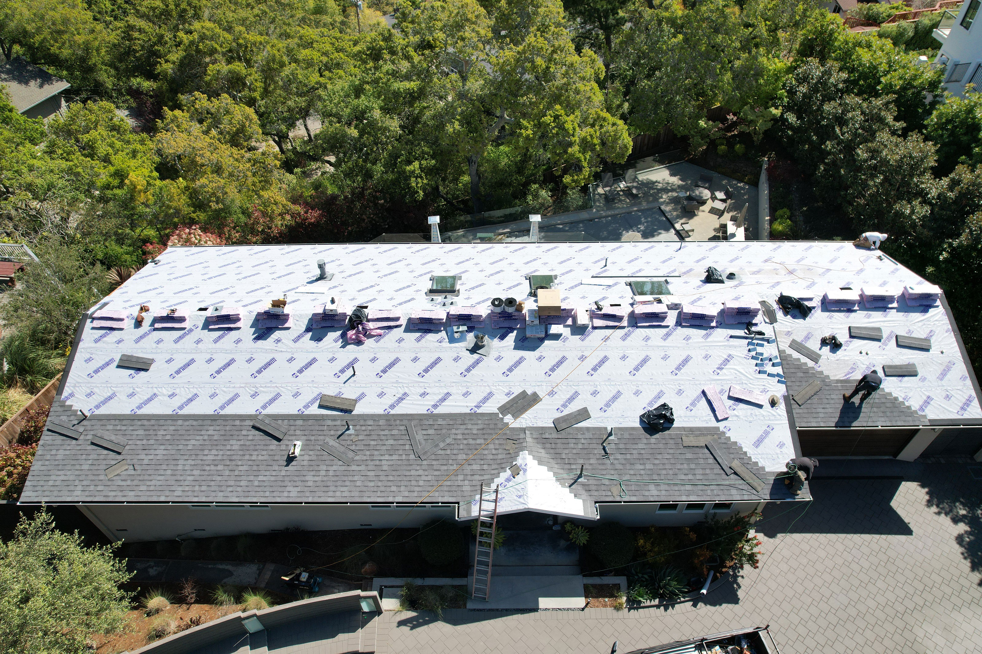The image shows a building with a visible roofing project underway, featuring blue tarps covering the roof surface, and there appears to be some construction or renovation work taking place on the exterior walls.