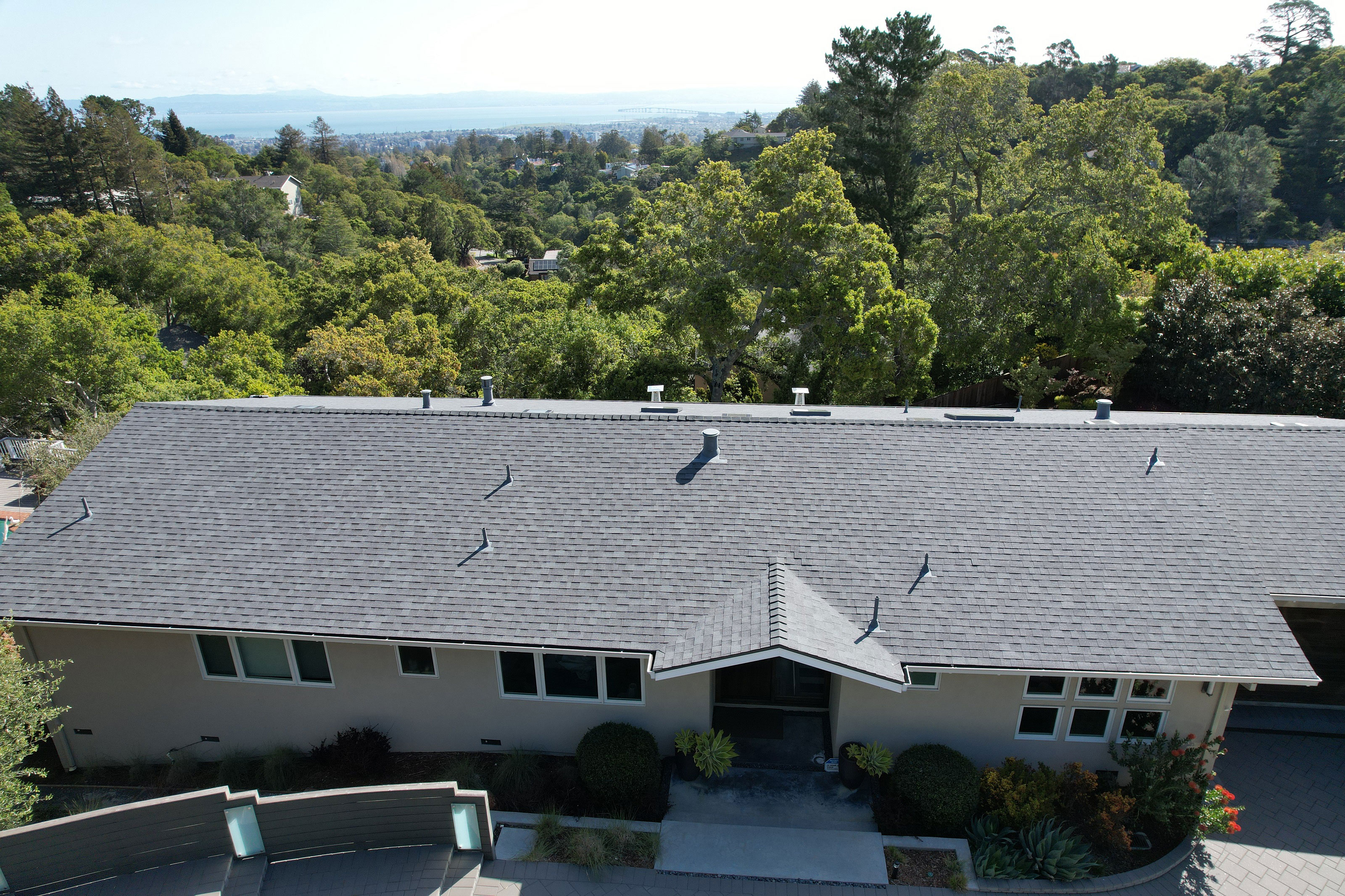 The image shows a two-story residential house with a gray shingle roof, a covered front porch, and a garage on the left side of the building. The house has multiple windows and is surrounded by landscaping.