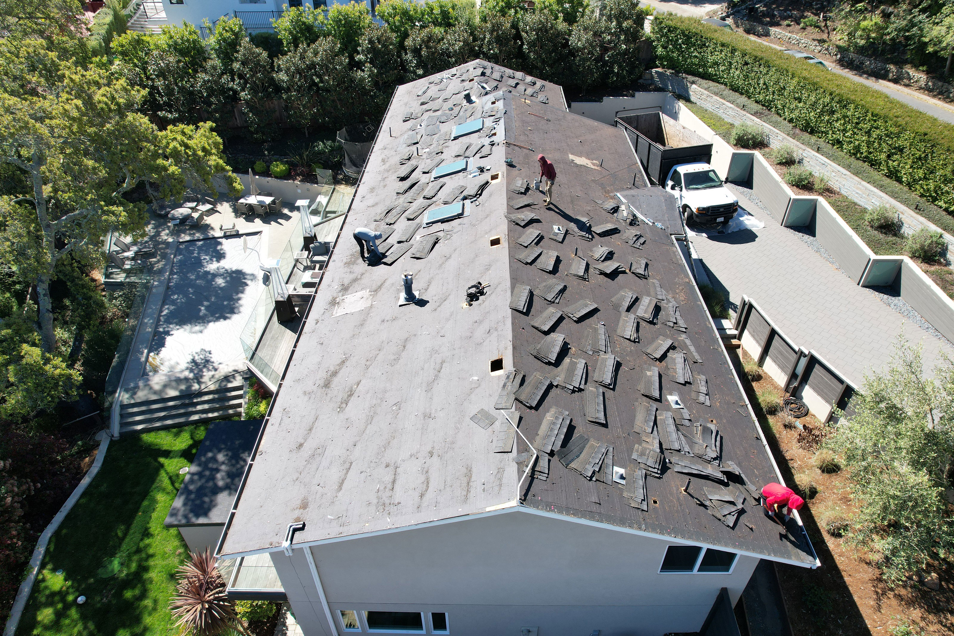 The image shows a house with a damaged roof under repair, featuring multiple openings and exposed plywood sheathing, and there are workers on the roof.