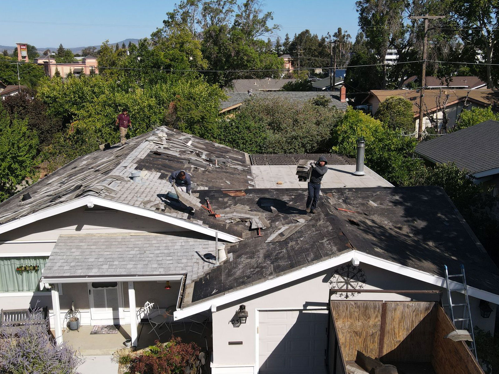 The image shows a residential property with a roof under repair. There are workers on the roof engaged in the renovation process, using tools such as hammers and nail guns. The house has a two-car garage, visible through the open door, and is surrounded by a neatly maintained yard with trees and shrubs. The sky is clear, suggesting it might be a sunny day.
