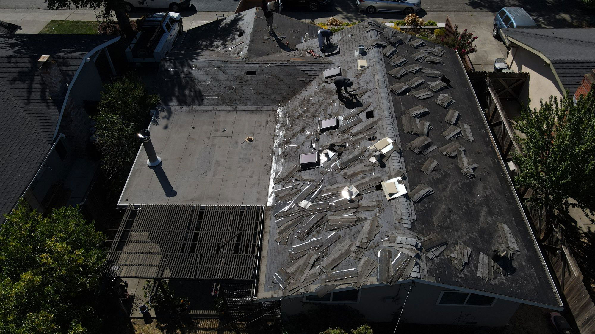 The image shows a house with a damaged roof under repair, featuring exposed rafters and shingles, taken from an elevated angle during daylight.