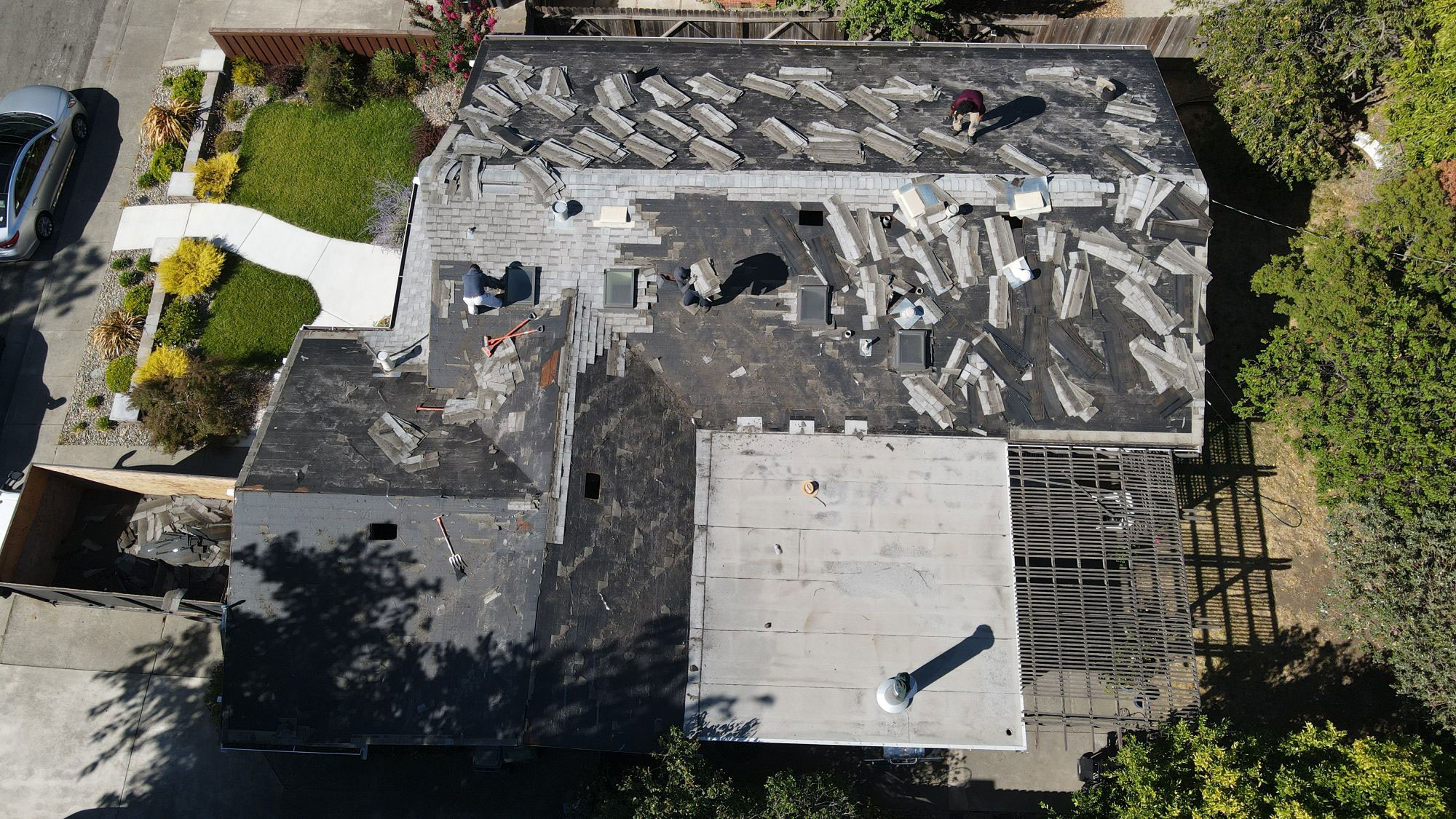 The image shows a residential building with an unfinished roof undergoing construction, with various materials scattered on the ground around it.