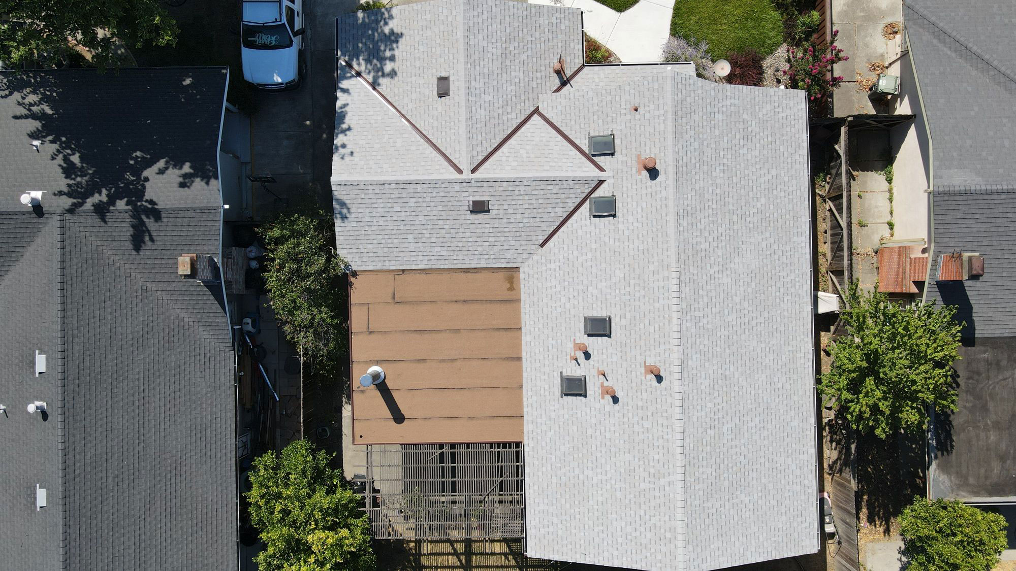 The image shows a residential house with a visible roofing project underway, featuring a patchwork of shingles in various stages of installation, set against a clear sky.