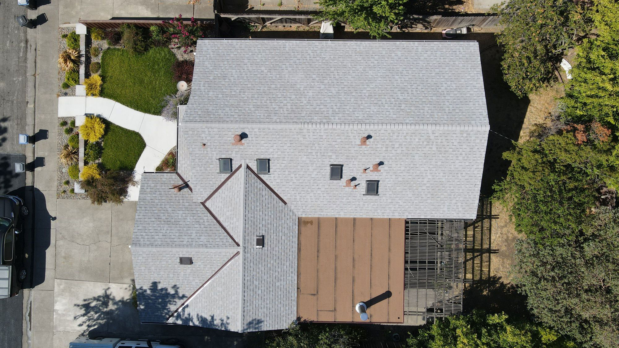 The image shows a two-story residential house with a gray roof, a brown deck, a white driveway, and a green lawn.
