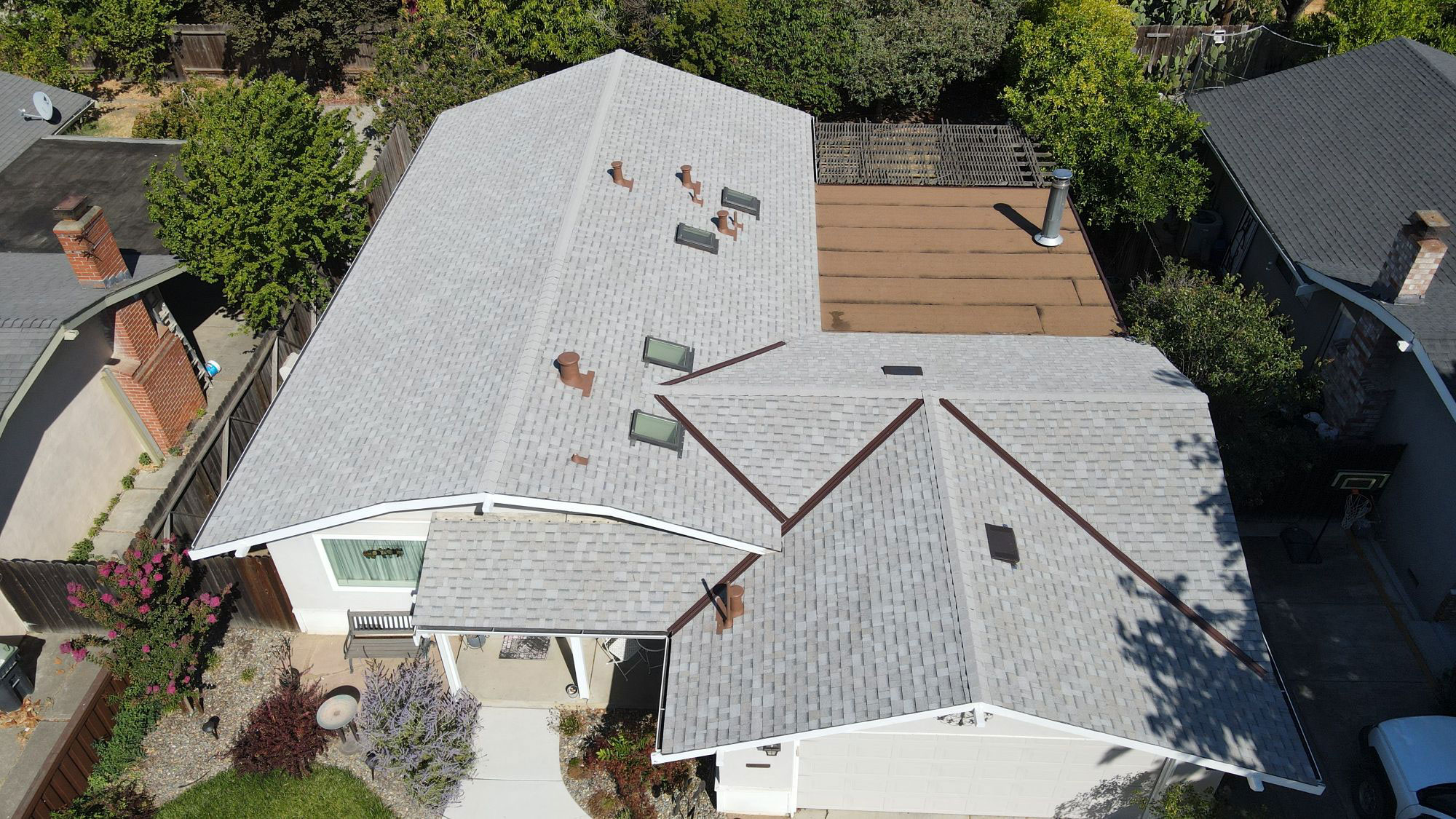 The image shows a residential house with a prominent gray roof featuring multiple skylights and solar panels, set against a backdrop of trees and a clear sky.