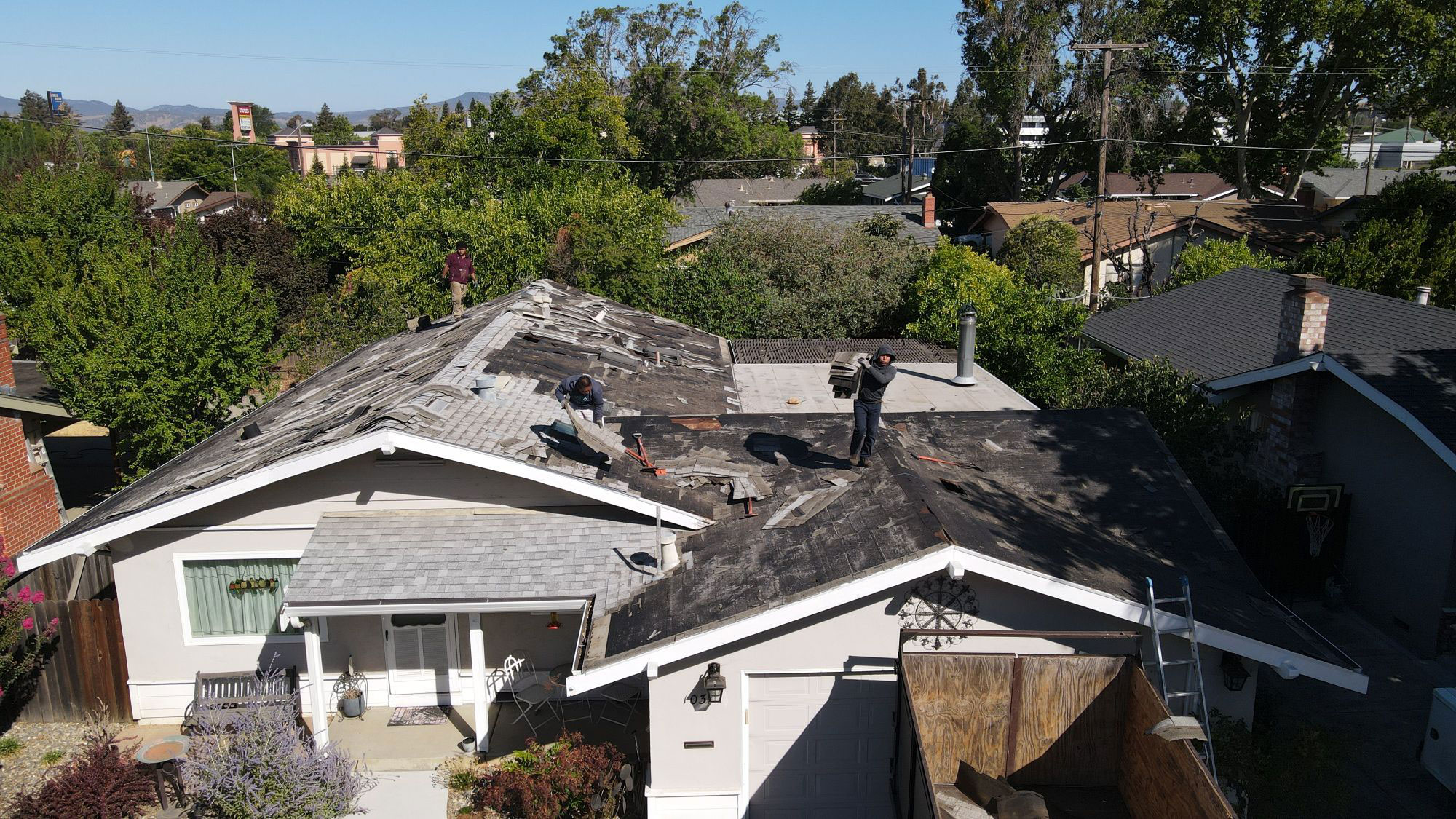 The image shows a residential area with a house undergoing roof repair work, featuring multiple people working on the project, and is taken from an elevated angle.