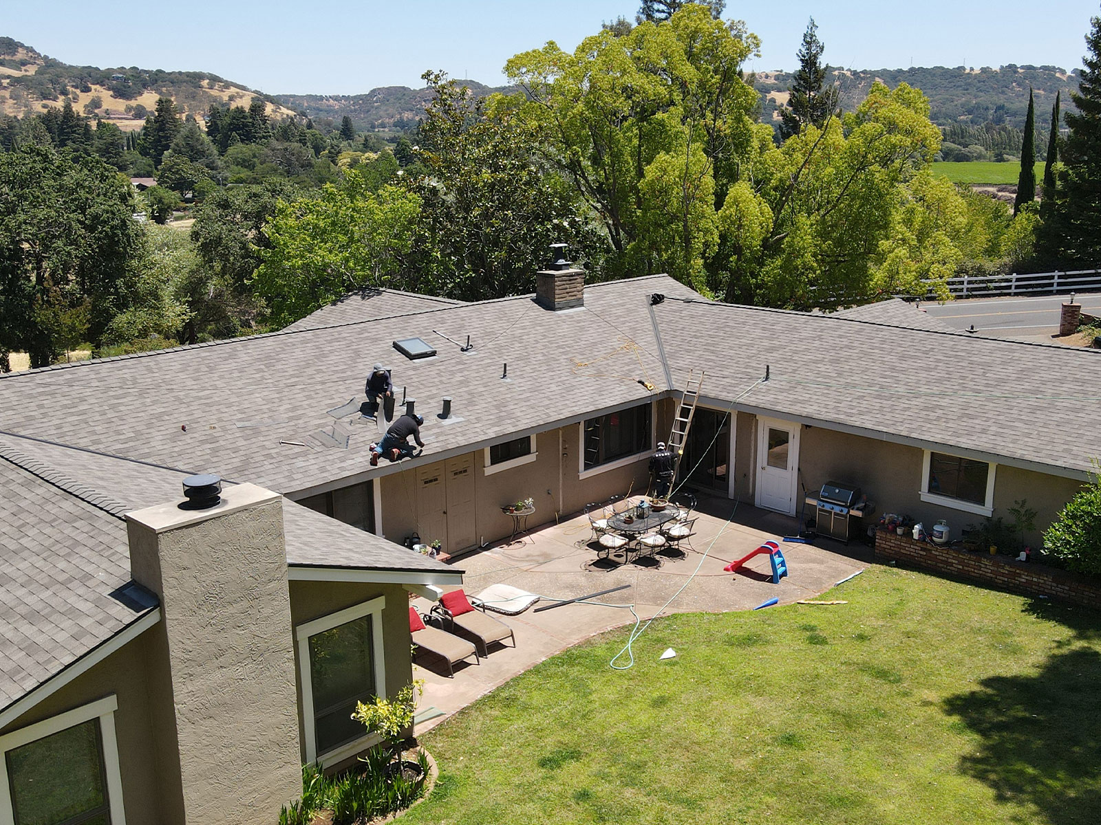 Aerial view of a residential property with a roof under construction, showcasing a yard, patio furniture, and a clear sky.