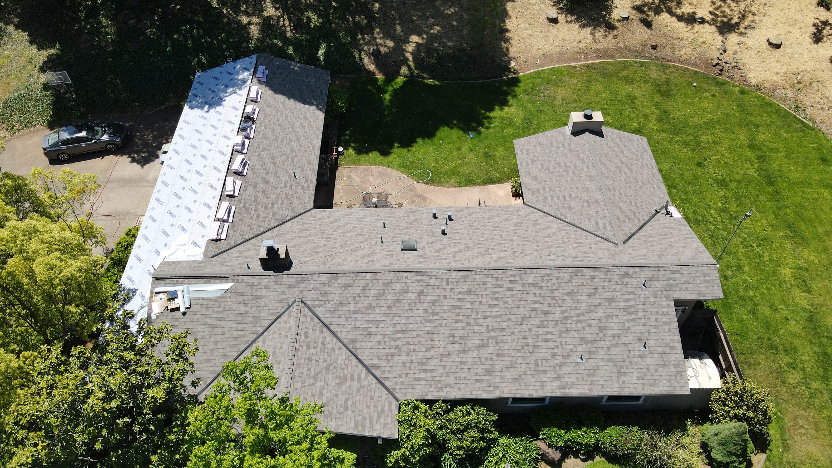 The image shows a two-story residential house with a shingled roof under construction, featuring a garage with an attached carport, set against a backdrop of trees and a clear sky.