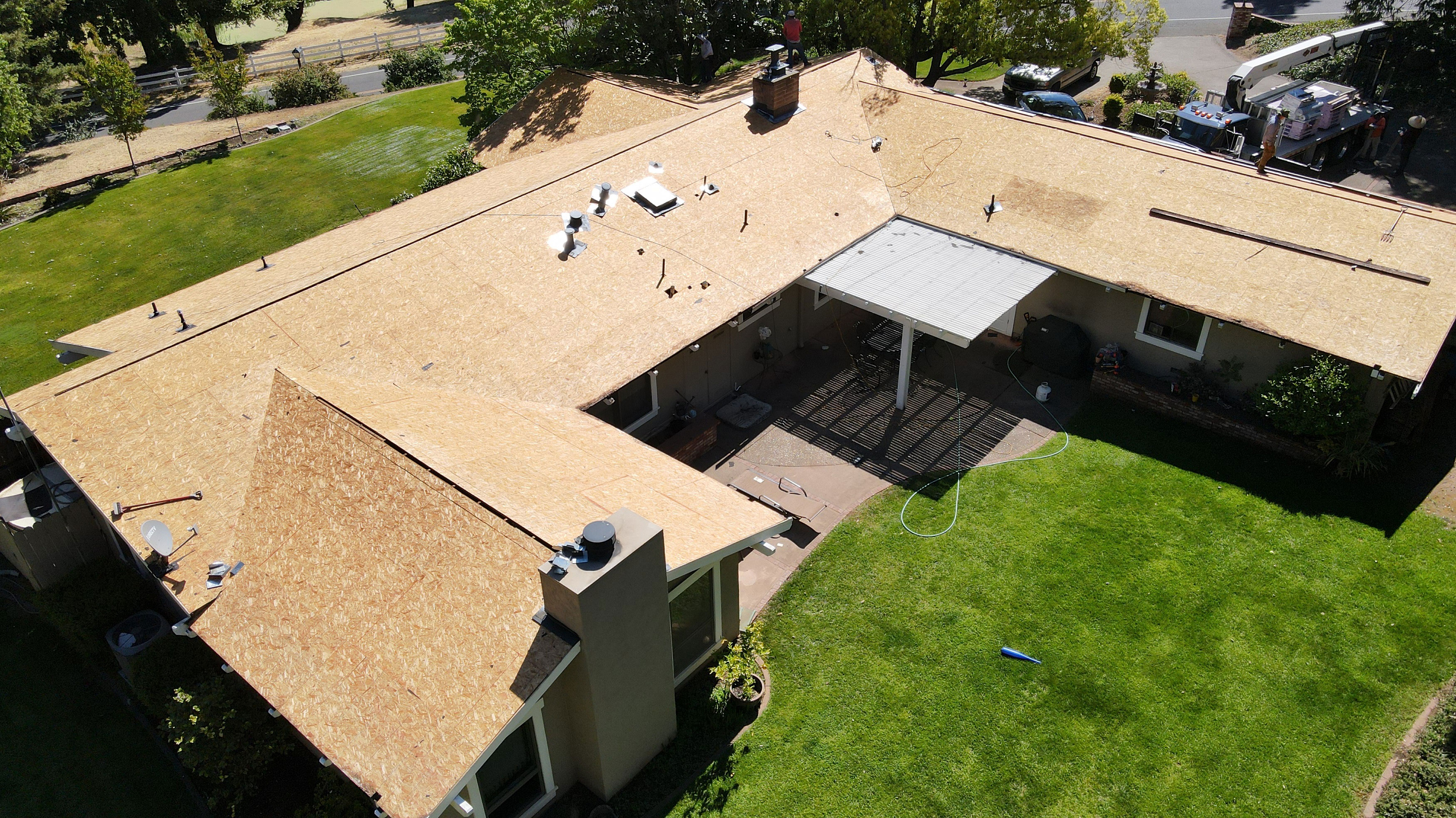 The image shows a residential house undergoing roofing work, with the old shingles removed and new ones being installed.