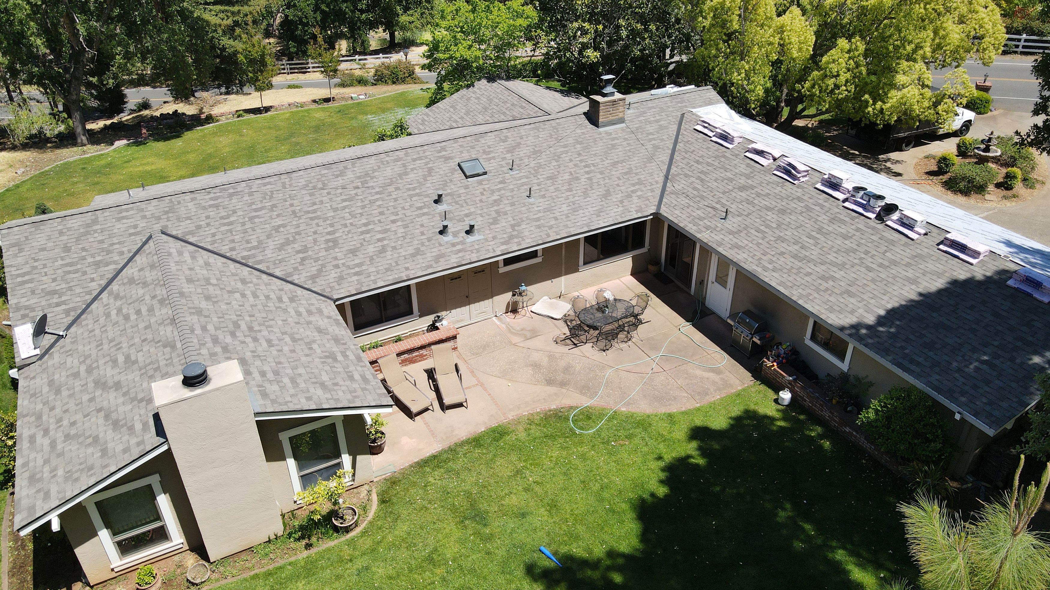 The image shows a single-story house with a shingled roof, a driveway, and a backyard area with a patio and lawn.