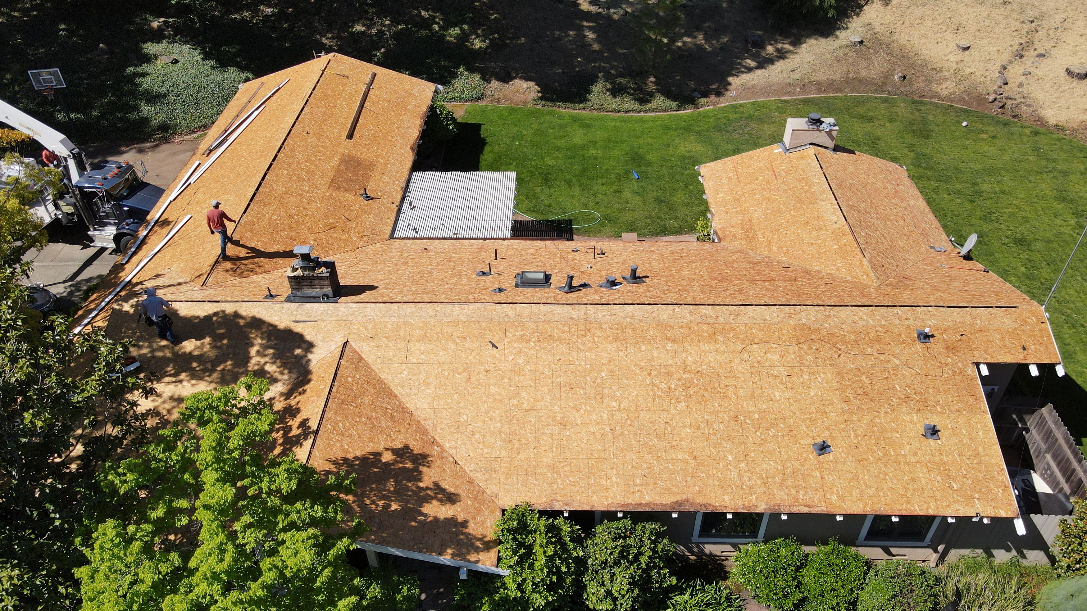 The image depicts a house under construction with a visible roofing process taking place, surrounded by a yard with trees and grass.