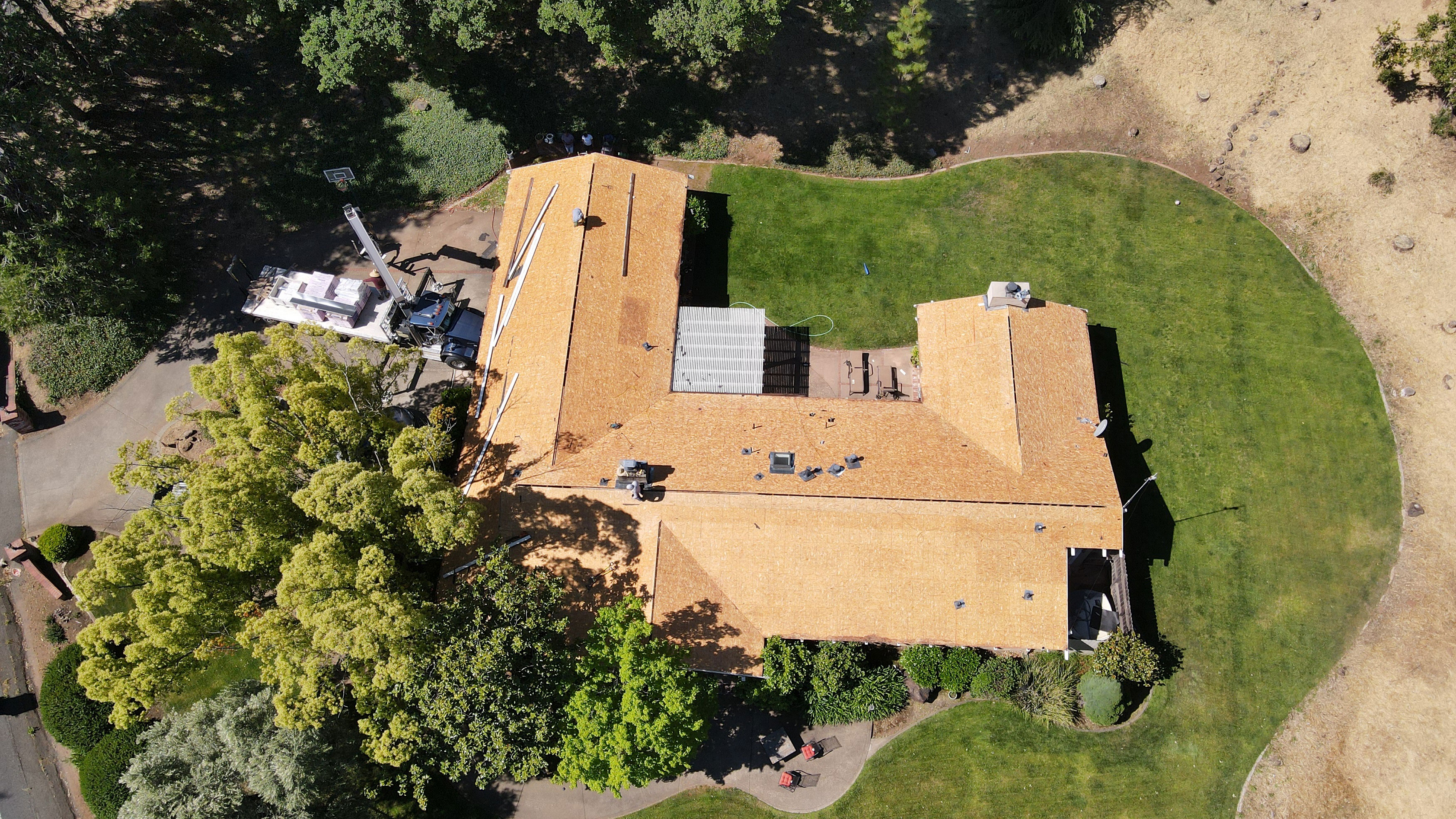 The image shows a large residential house with a flat roof under construction, surrounded by trees and a grassy lawn, with a clear sky above.