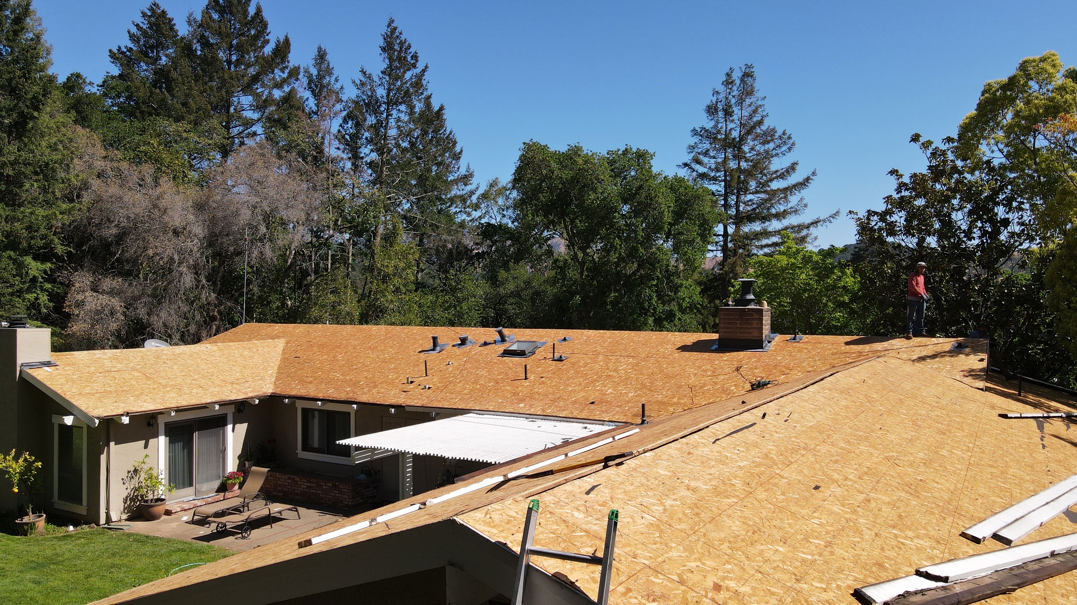 The image shows a residential property undergoing roof construction, with a partially completed structure visible against a clear sky and greenery.