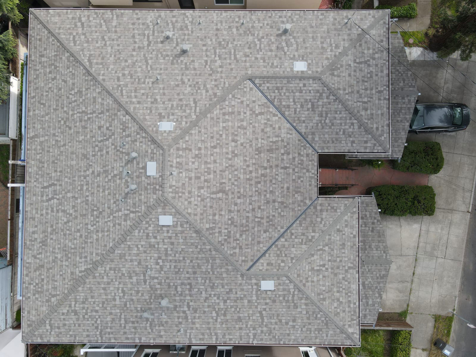 The image shows a residential house with a flat roof, featuring interlocking shingles and visible gutter systems, situated on a street with houses on both sides and a clear sky above.
