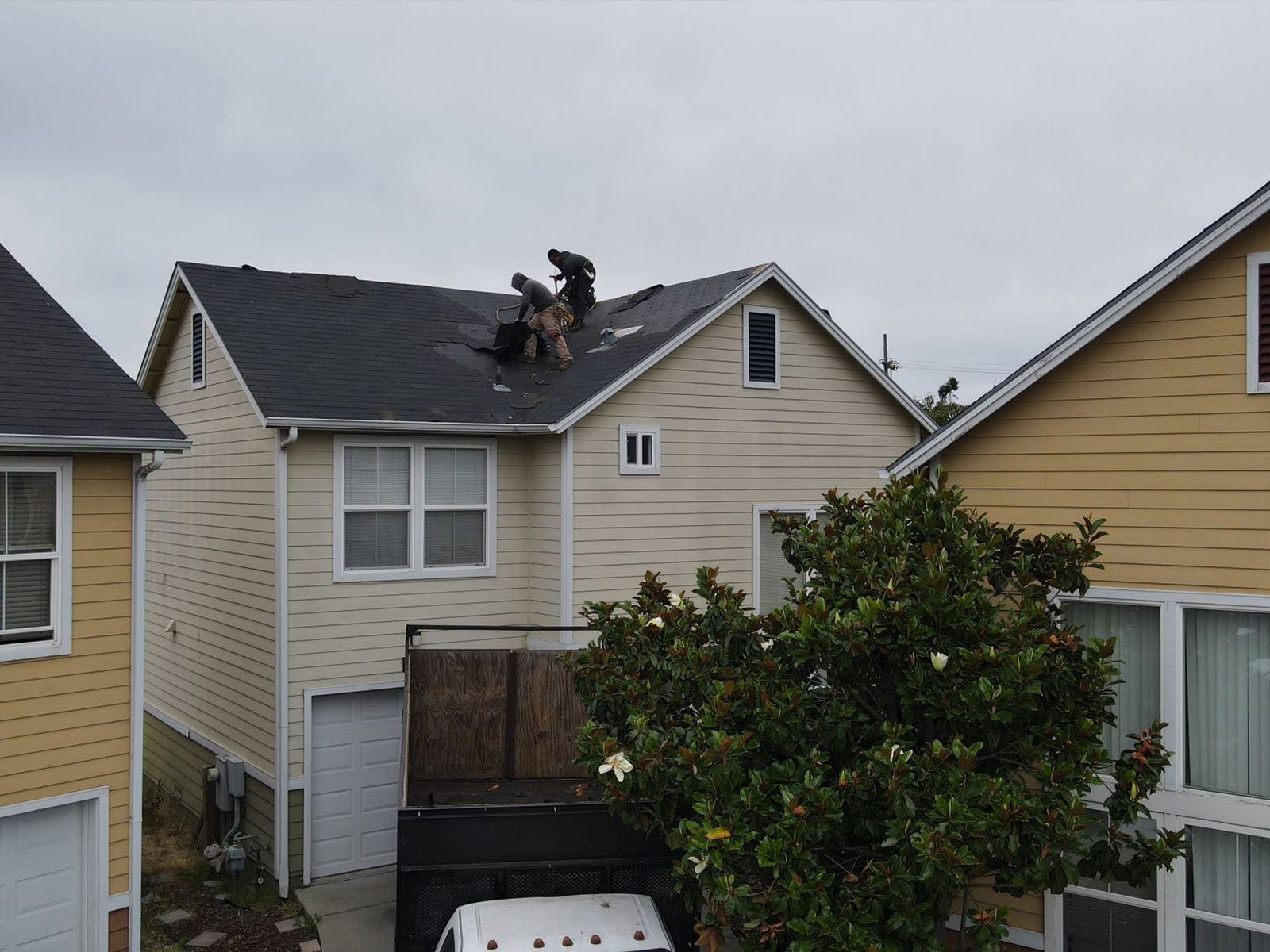 The image shows a residential house with a damaged roof under repair, featuring workers on the roof and a truck parked nearby.