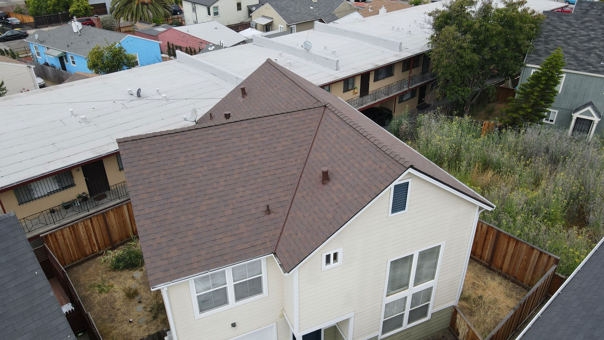 The image shows a two-story residential house with a dark brown roof, situated within a suburban neighborhood with other houses visible in the background.