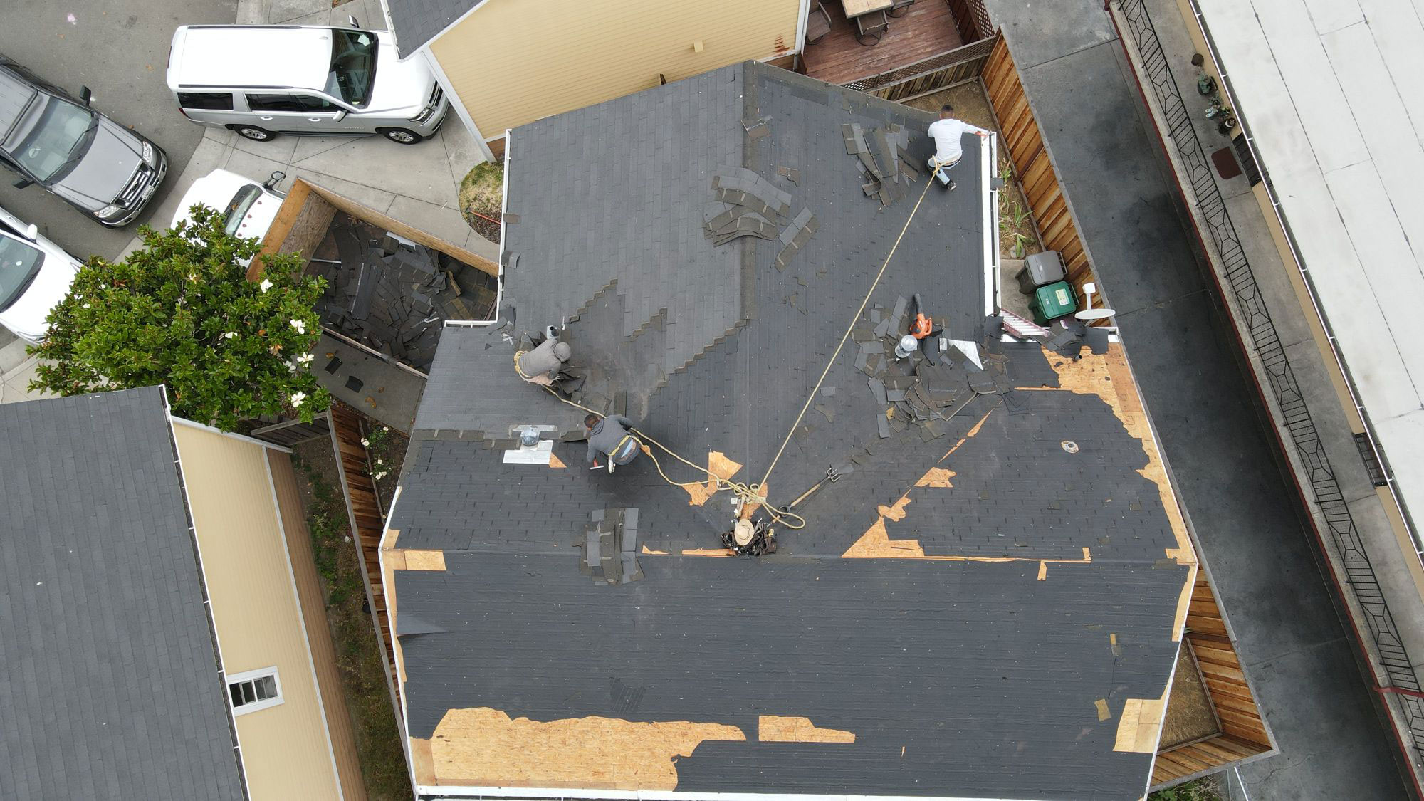 The image shows a residential house with a damaged roof under repair, featuring visible tarps and exposed insulation, amidst ongoing construction work on an adjacent building.