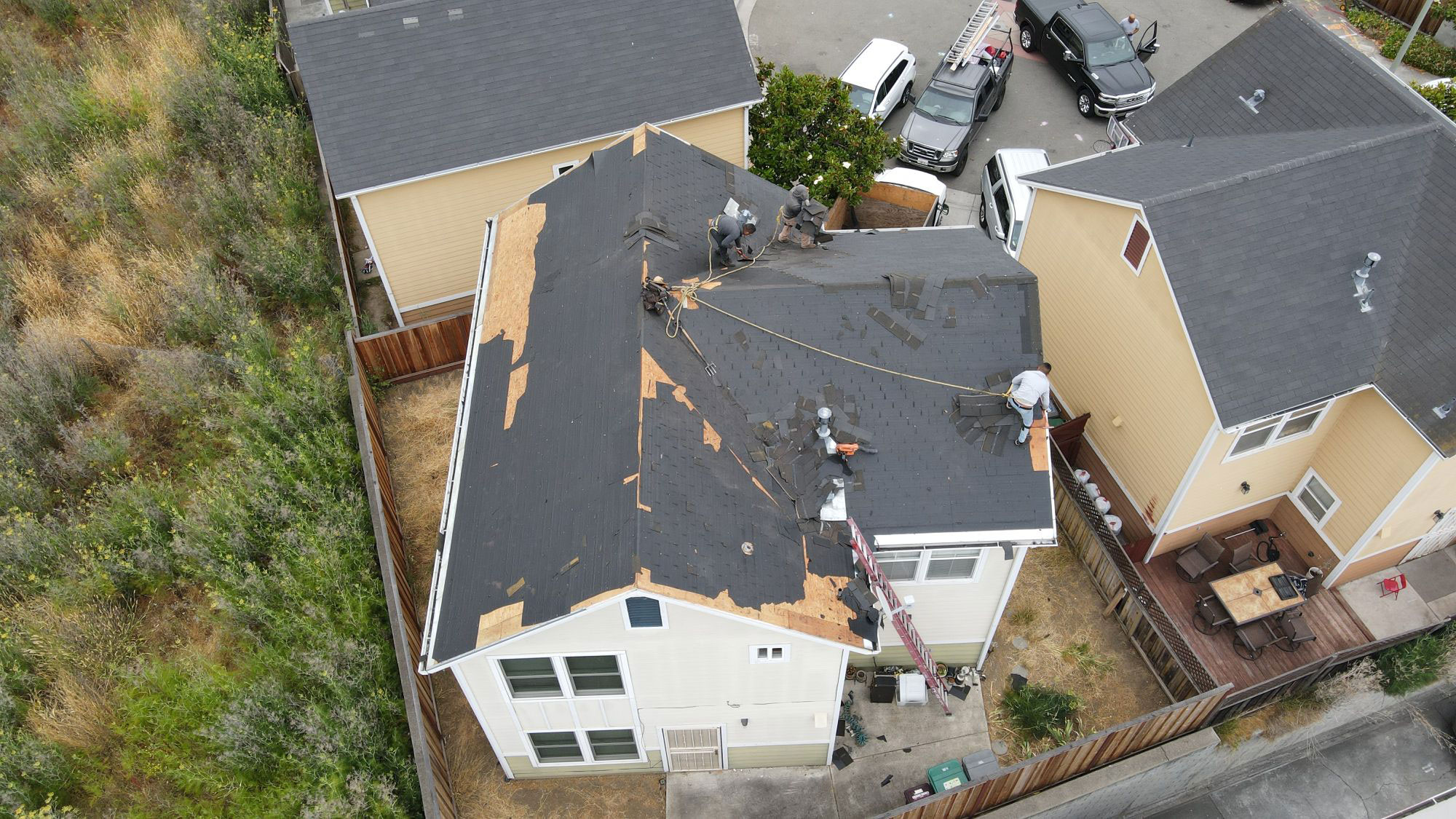 The image shows a residential house with a damaged roof under repair, with workers on the roof and surrounding areas, and vehicles parked nearby.