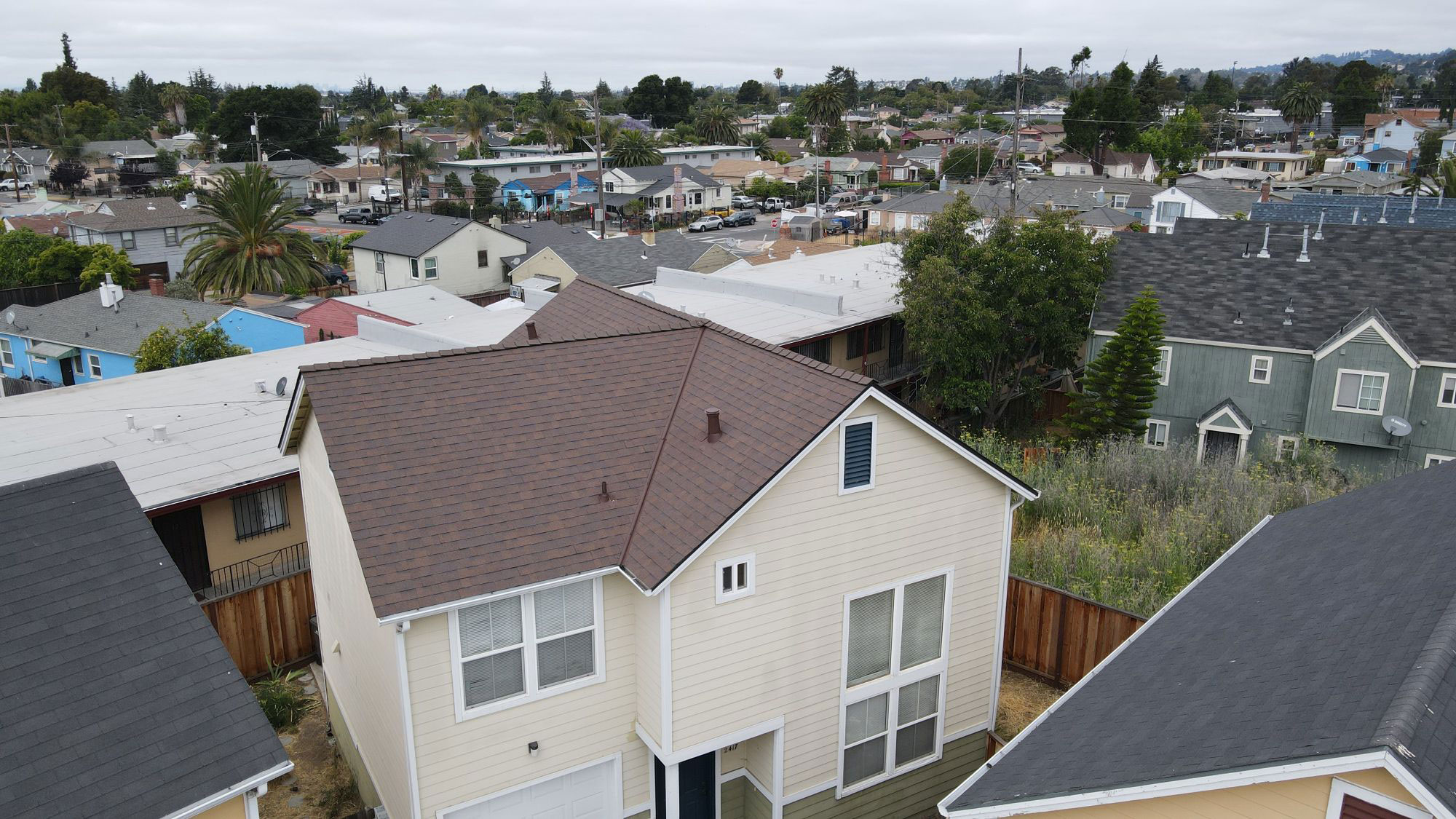 The image shows a residential house with a shingled roof, set against a clear sky. It appears to be taken from an elevated angle, possibly from another house across the street. The house has a white exterior and is surrounded by other homes in a suburban setting.