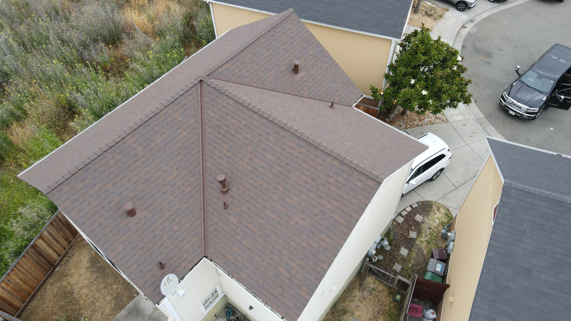 The image shows a residential house with a brown shingle roof, viewed from an angle that captures its front facade and part of the surrounding landscape.