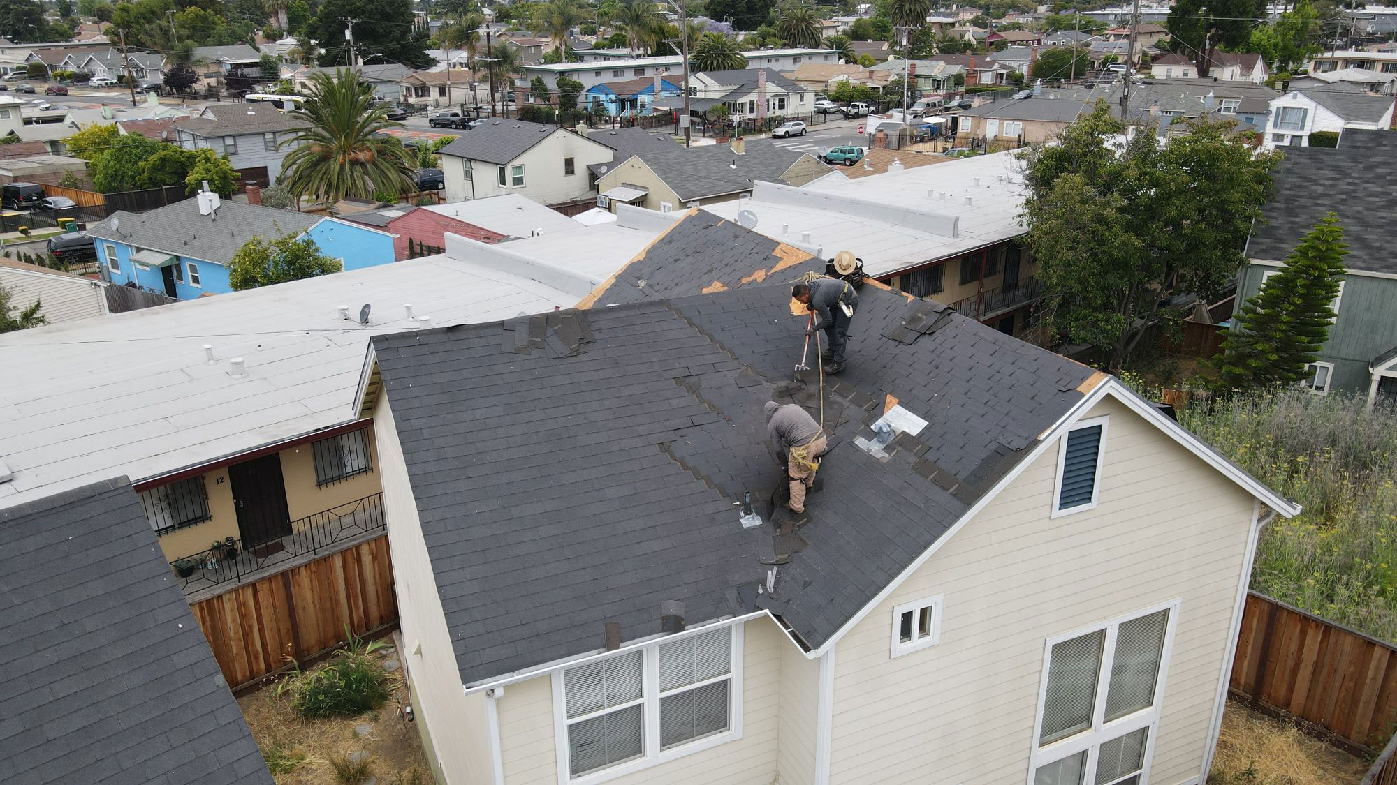The image shows a residential area with a house undergoing roof repair, with visible tarps on the roof and a person working on the structure.