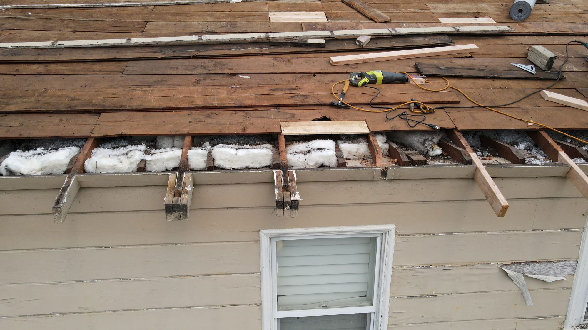 The image shows a roof under construction with visible wooden planks and exposed insulation, taken from an exterior perspective showing part of a house.