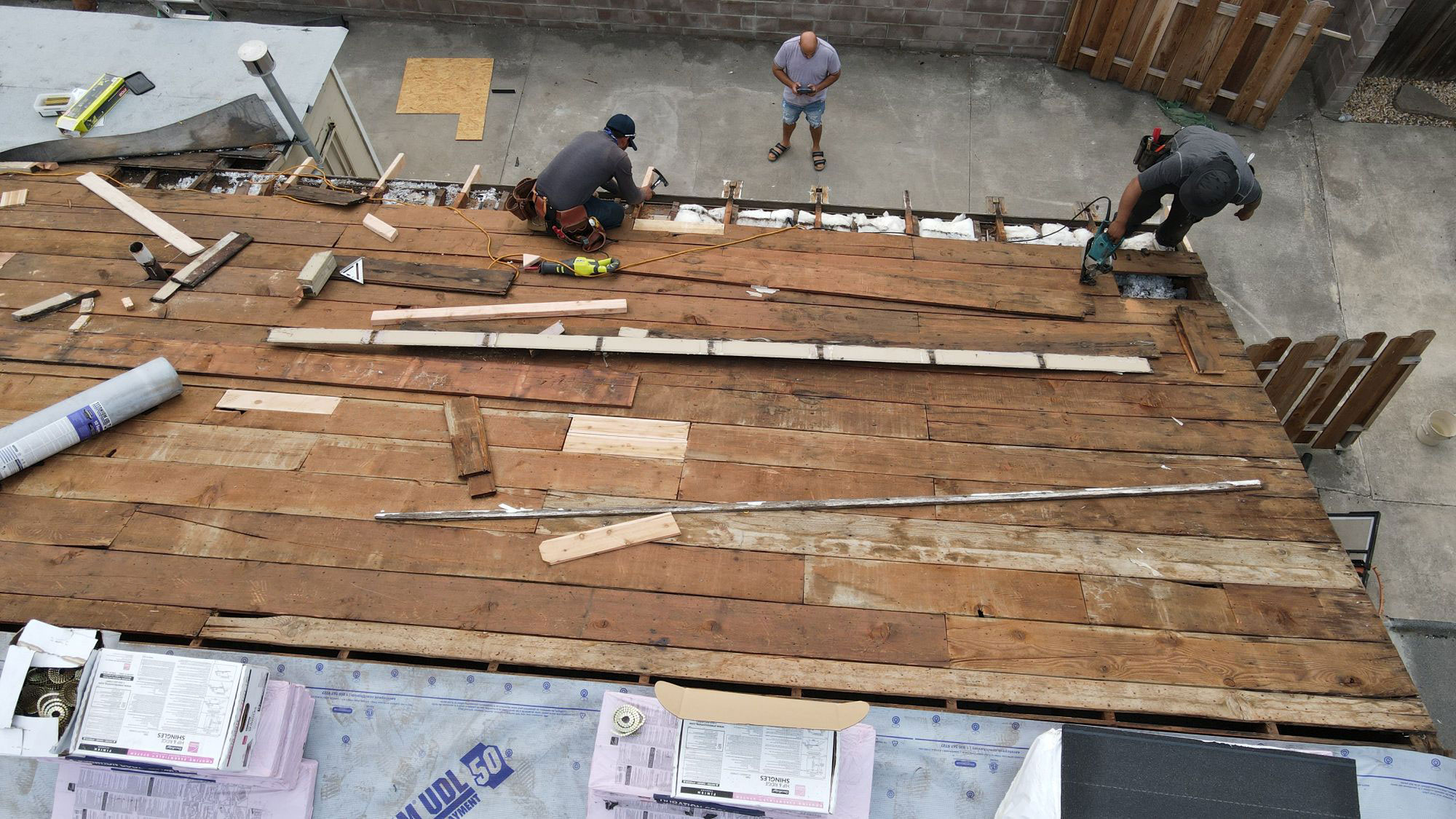The image shows construction work being done on a roof with wooden planks visible, workers on site, and various tools and materials present.