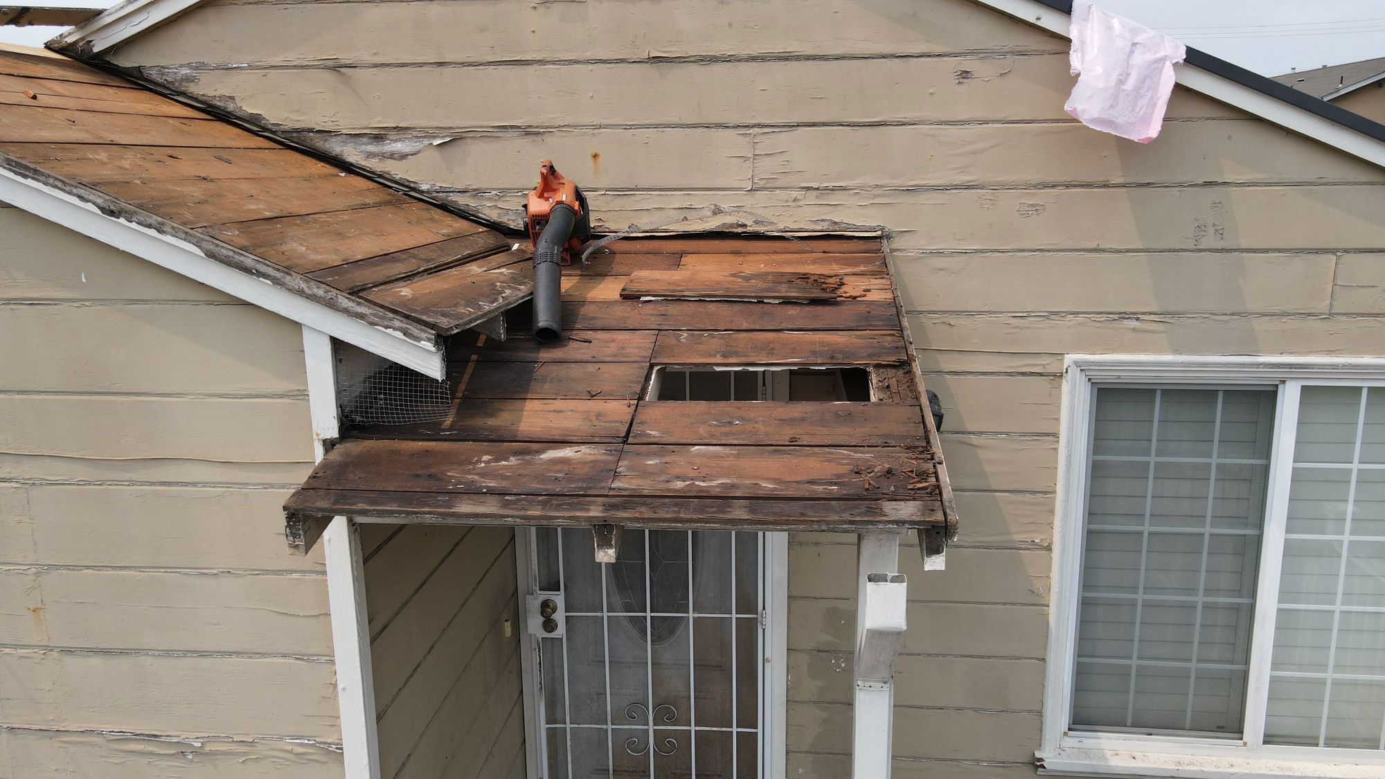 The image shows a house with a roof that appears to be damaged or under repair, featuring visible wood planks and a person sitting on the edge of the roof.