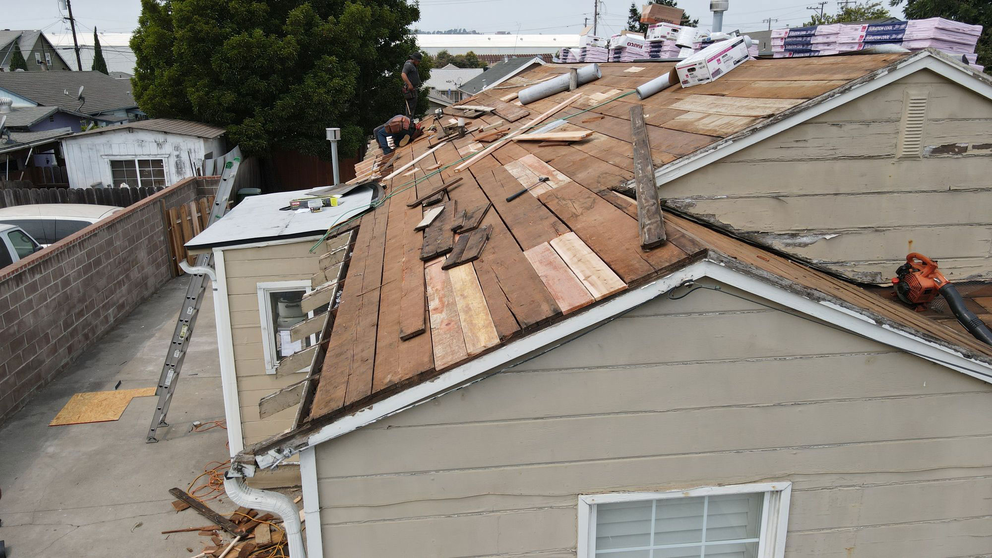 The image shows a house with a damaged roof under repair, featuring visible wooden planks and construction materials on the ground.