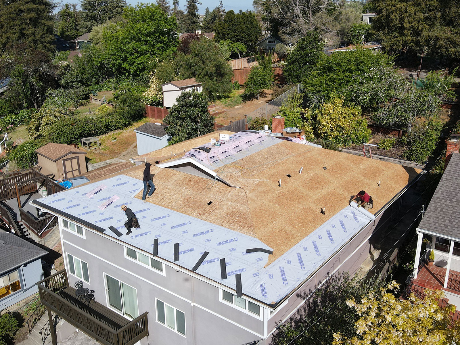 The image shows a residential house undergoing roof construction with workers on the scaffolding, surrounded by a fenced yard with a garden area and trees.