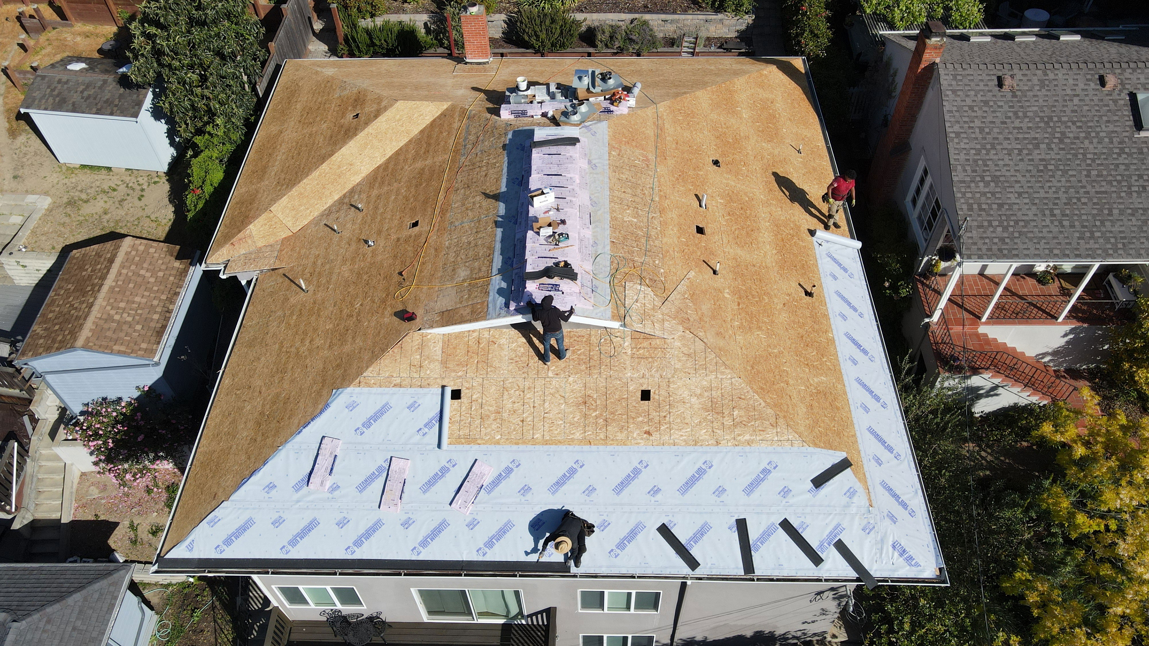 The image depicts a residential house under construction with a partially completed roof and workers on site, set against a clear sky and surrounded by a fence.