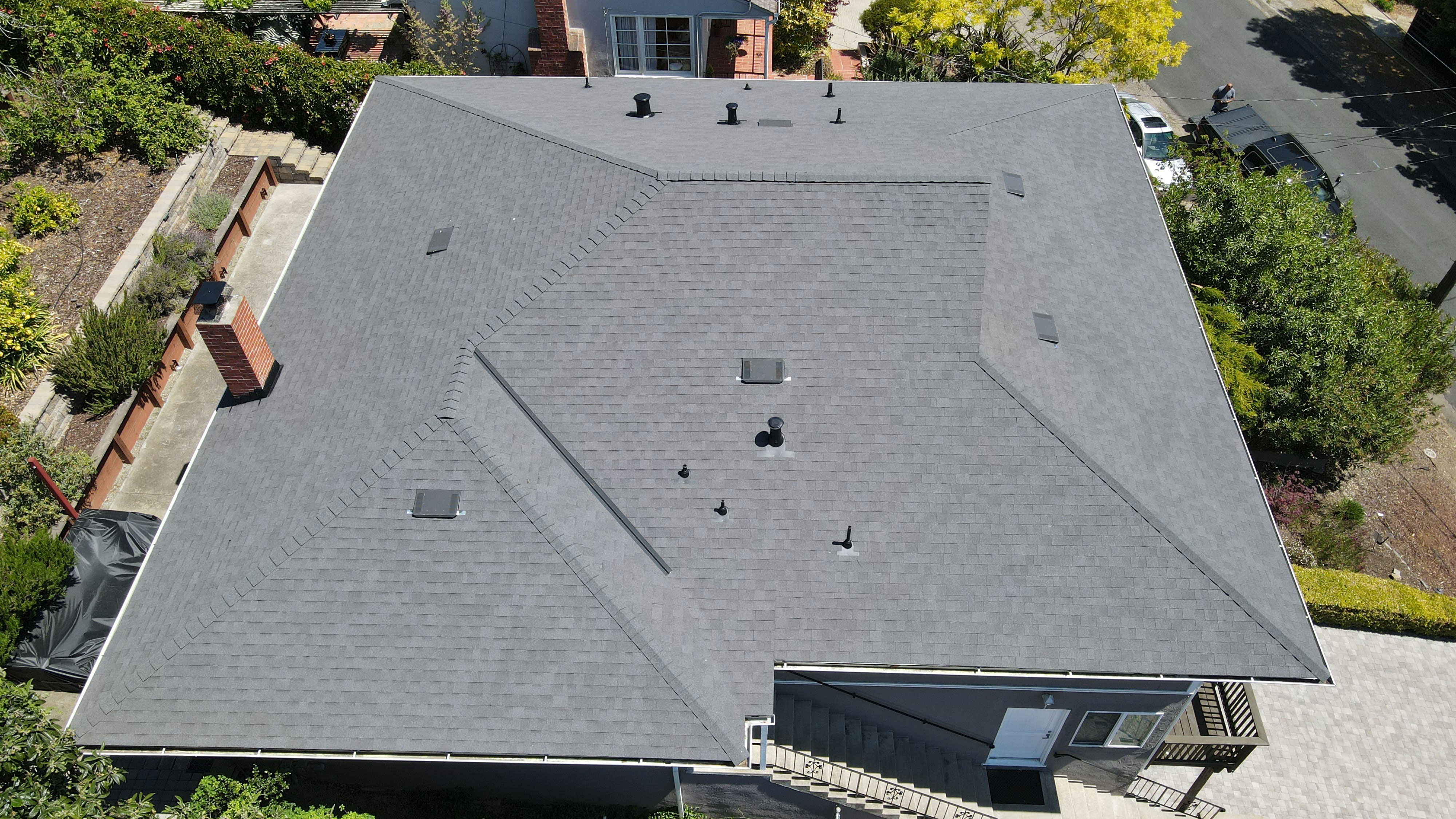 A photograph of a single-story house with a dark gray shingled roof under construction, viewed from an angle, showing the front facade.