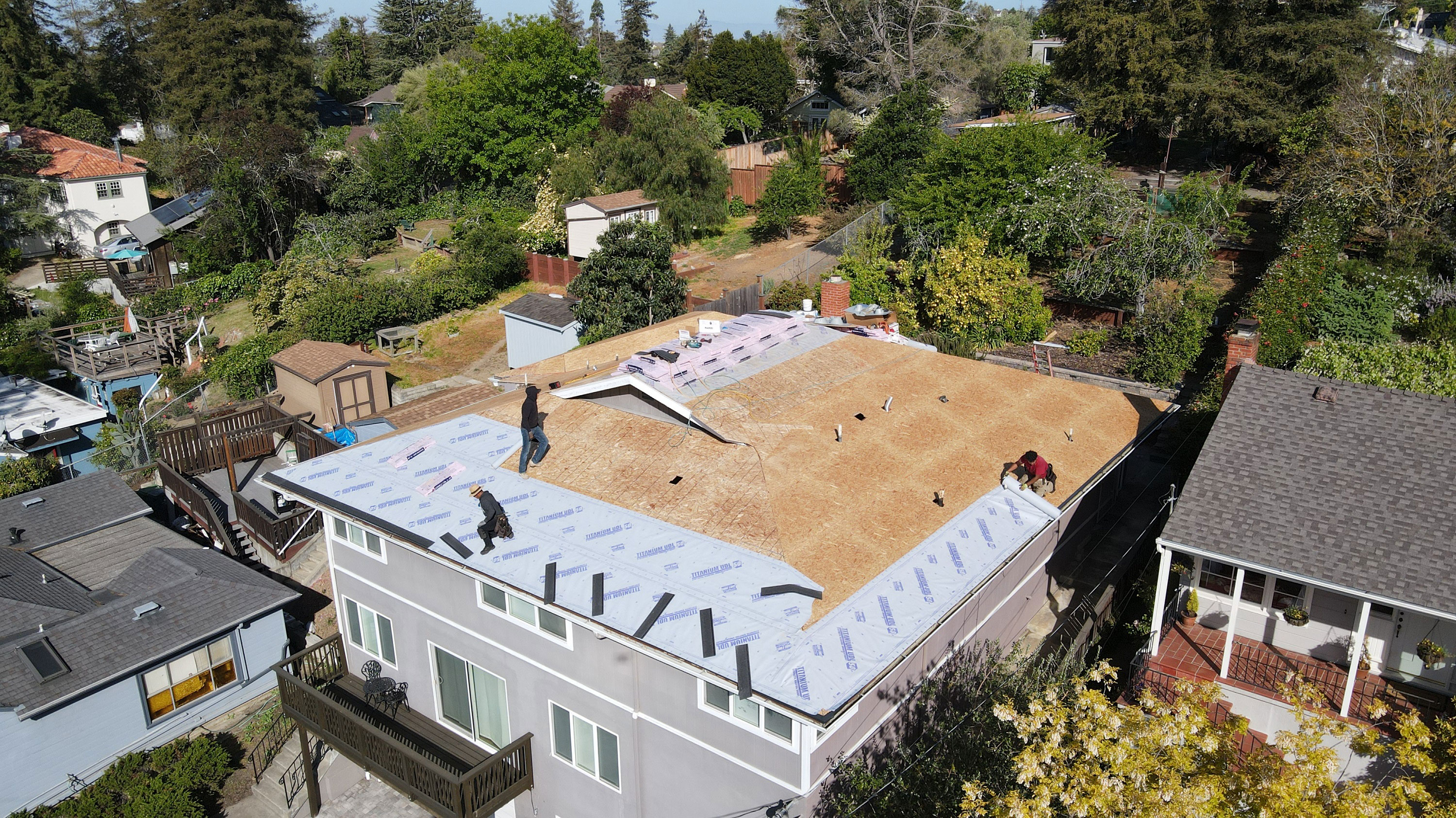 A house under construction with a roof partially covered in plywood, surrounded by residential houses on a sunny day.