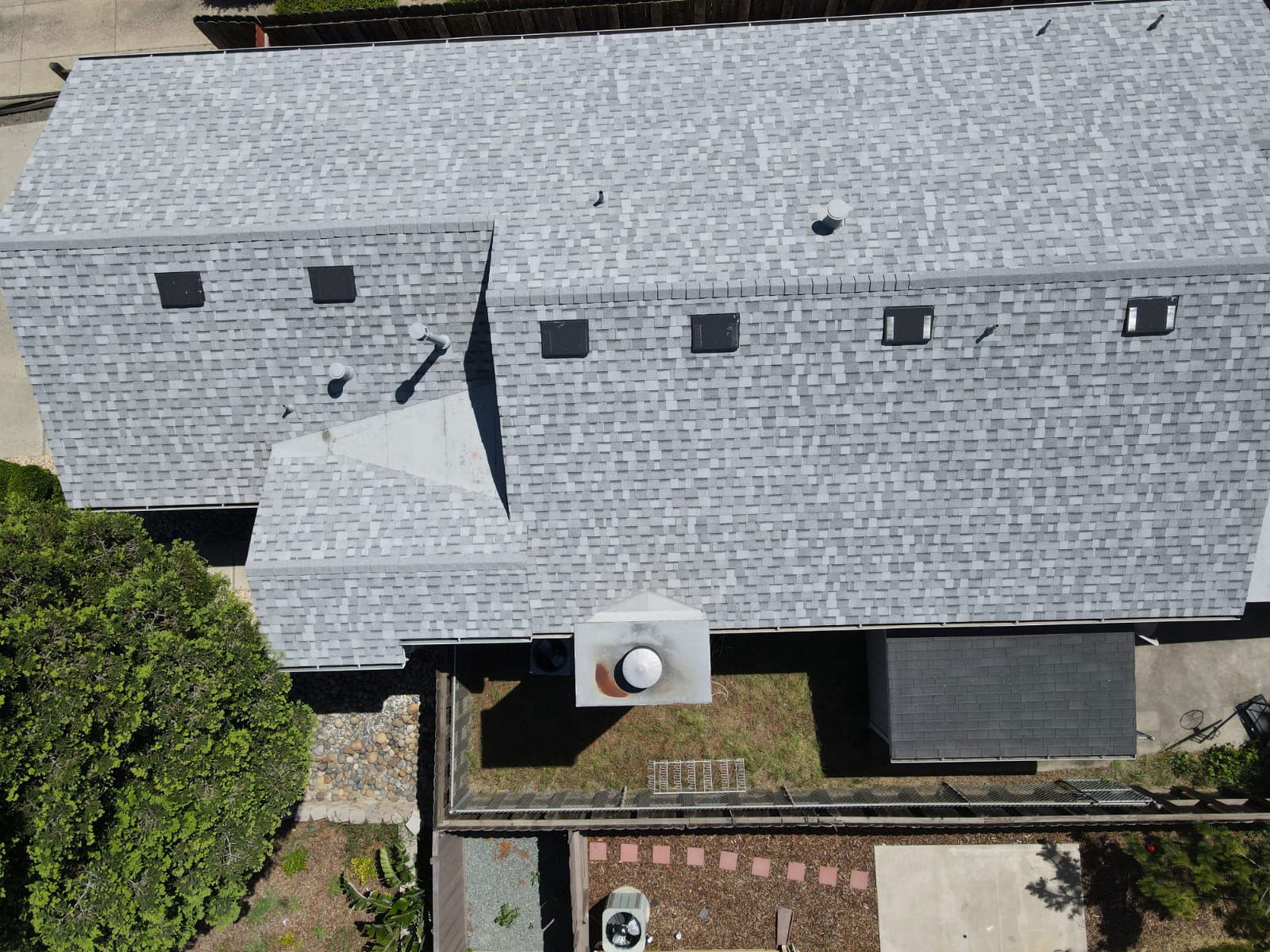 The image depicts a two-story building with a shingled roof, featuring a prominent sloped section on the left side and a flat roof on top of the main structure. There are multiple windows visible, some with shutters, and a single visible door. A satellite dish is mounted on the roof, and there s a chimney stack on the right side. The building appears to be constructed from concrete block or similar material, and it has a grayish color scheme. The surrounding area includes a paved driveway and a section of grass with some shrubbery. There are no visible people or moving vehicles in the image.