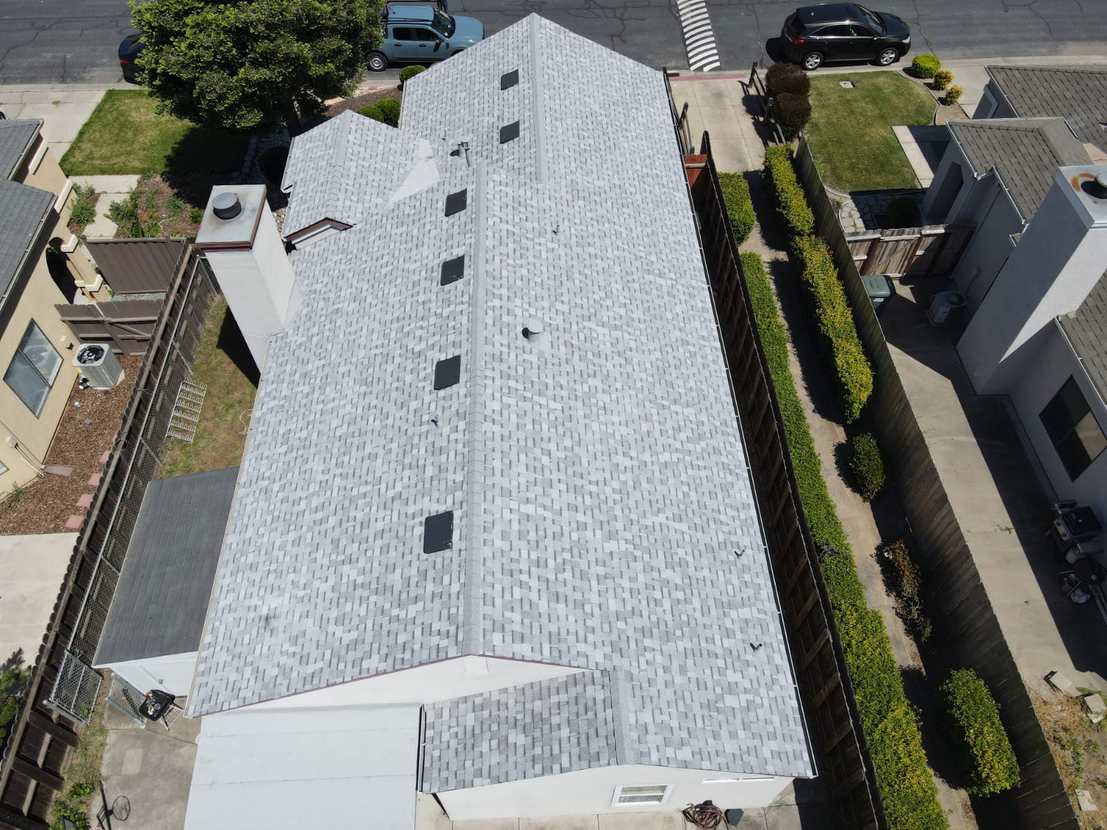 The image depicts a single-story residential building with a gray metal roof, featuring a series of skylights along its length, situated within a suburban neighborhood with houses on either side.