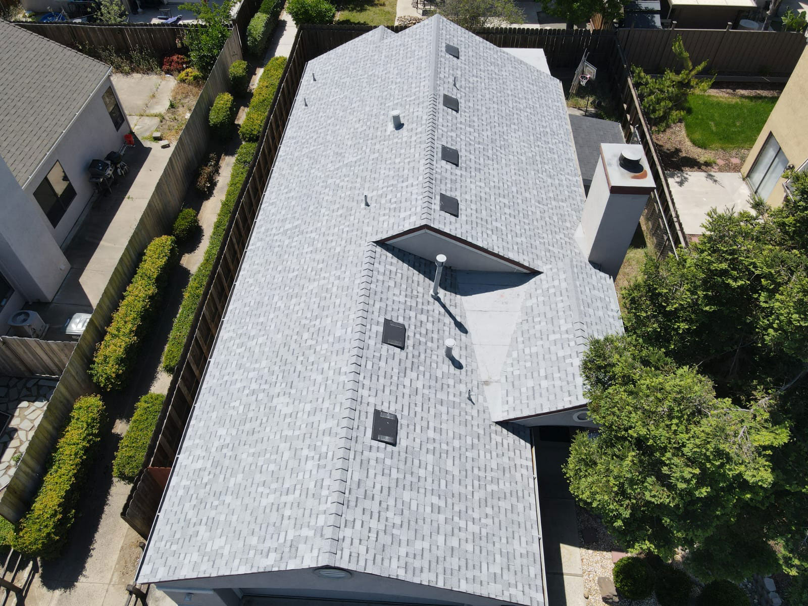 This is a photograph of a single-story residential house with a grey shingle roof, featuring multiple skylights and a prominent front gable with a triangular shape. The property has a fenced backyard and is situated in a suburban neighborhood.