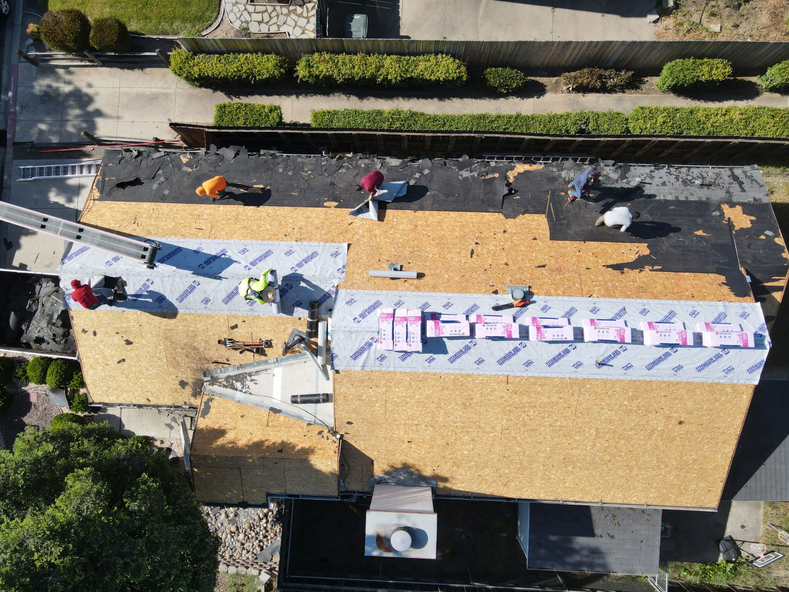 The image shows a roof under construction with workers on site, surrounded by a wooden fence and some landscaping features.