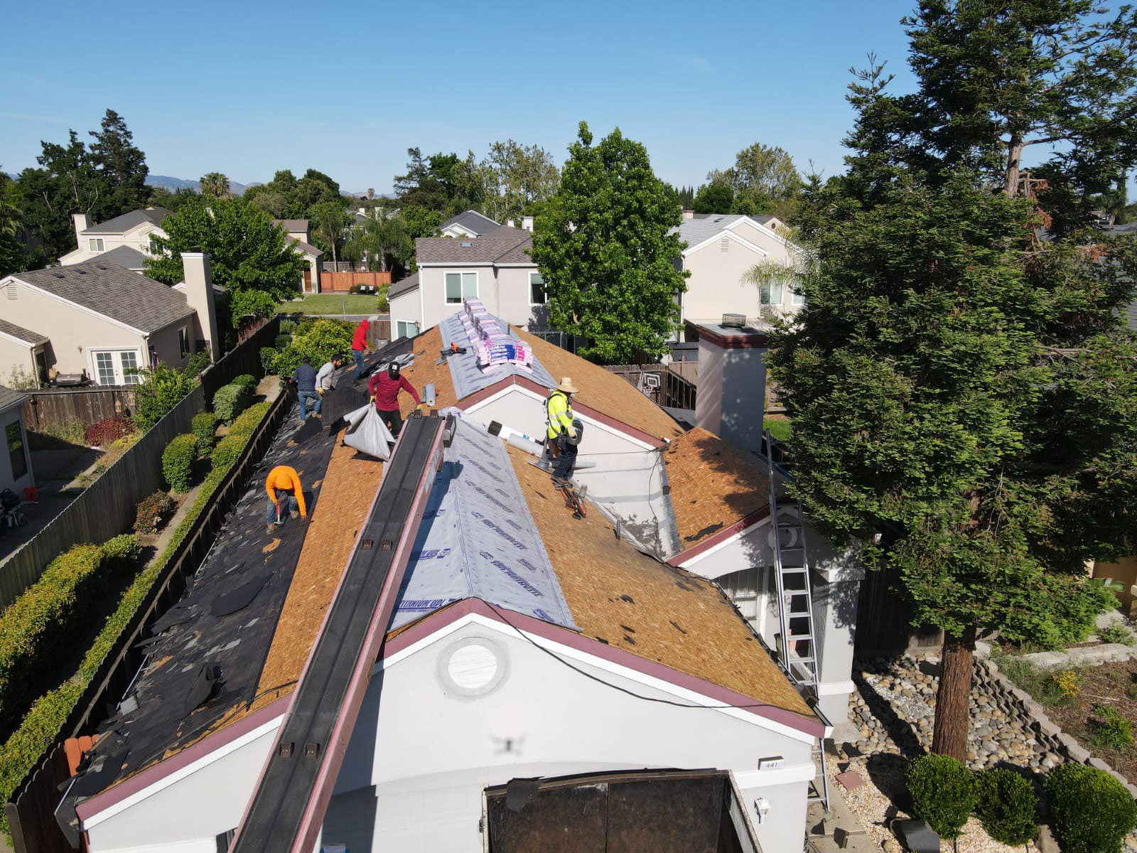 The image shows a residential property with a roof under construction or repair, featuring workers on the roof and scaffolding around the structure.