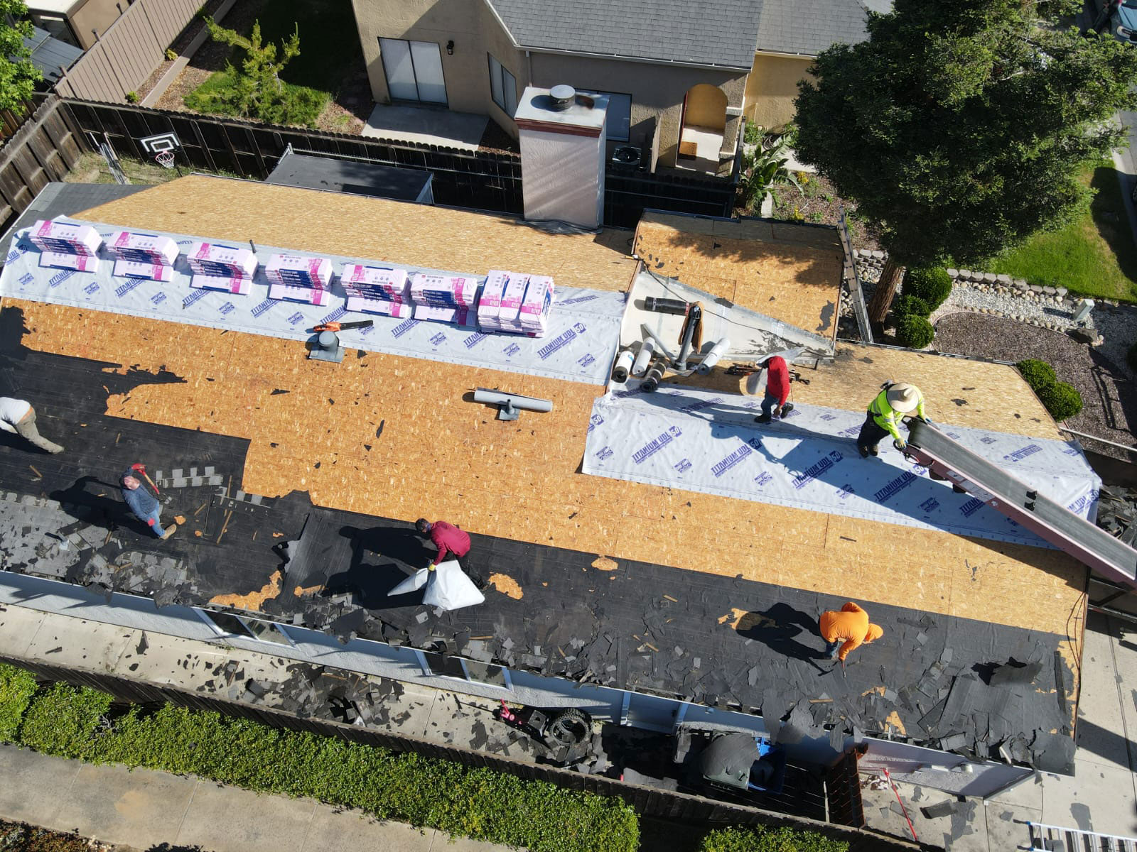 The image shows a roofing construction site with workers on top of a partially completed roof, surrounded by building materials and tools.