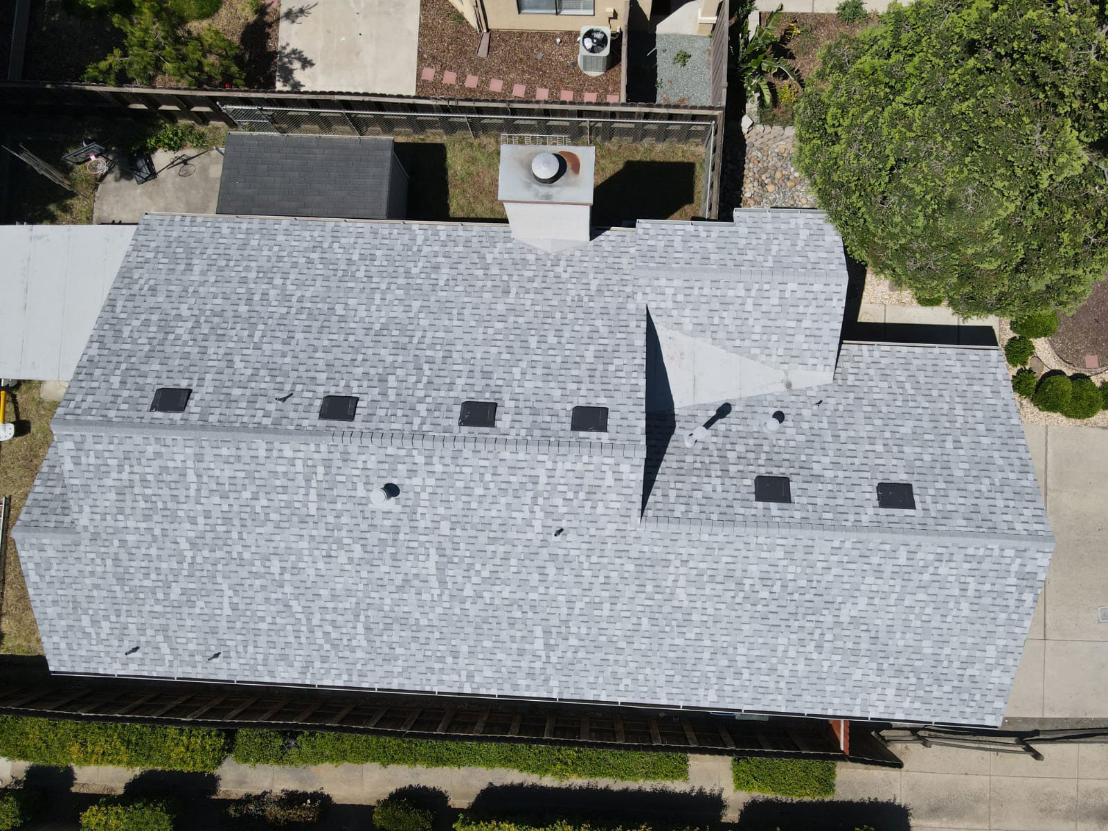The image shows a building with a gray shingle roof, featuring multiple skylights, set against a background of trees and a clear sky.