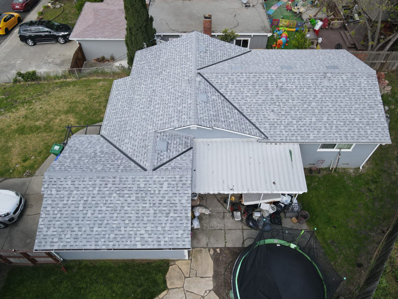 The image shows a house with a gray shingle roof, featuring a peaked design with multiple slopes, situated in a suburban neighborhood with visible cars parked on the street behind the property.