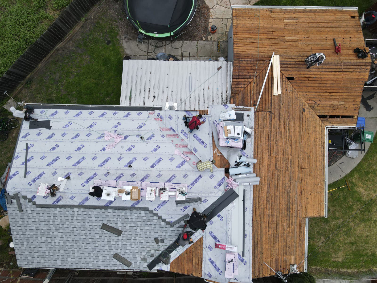 The image shows a house under construction with visible scaffolding, roofing materials, and workers on-site.
