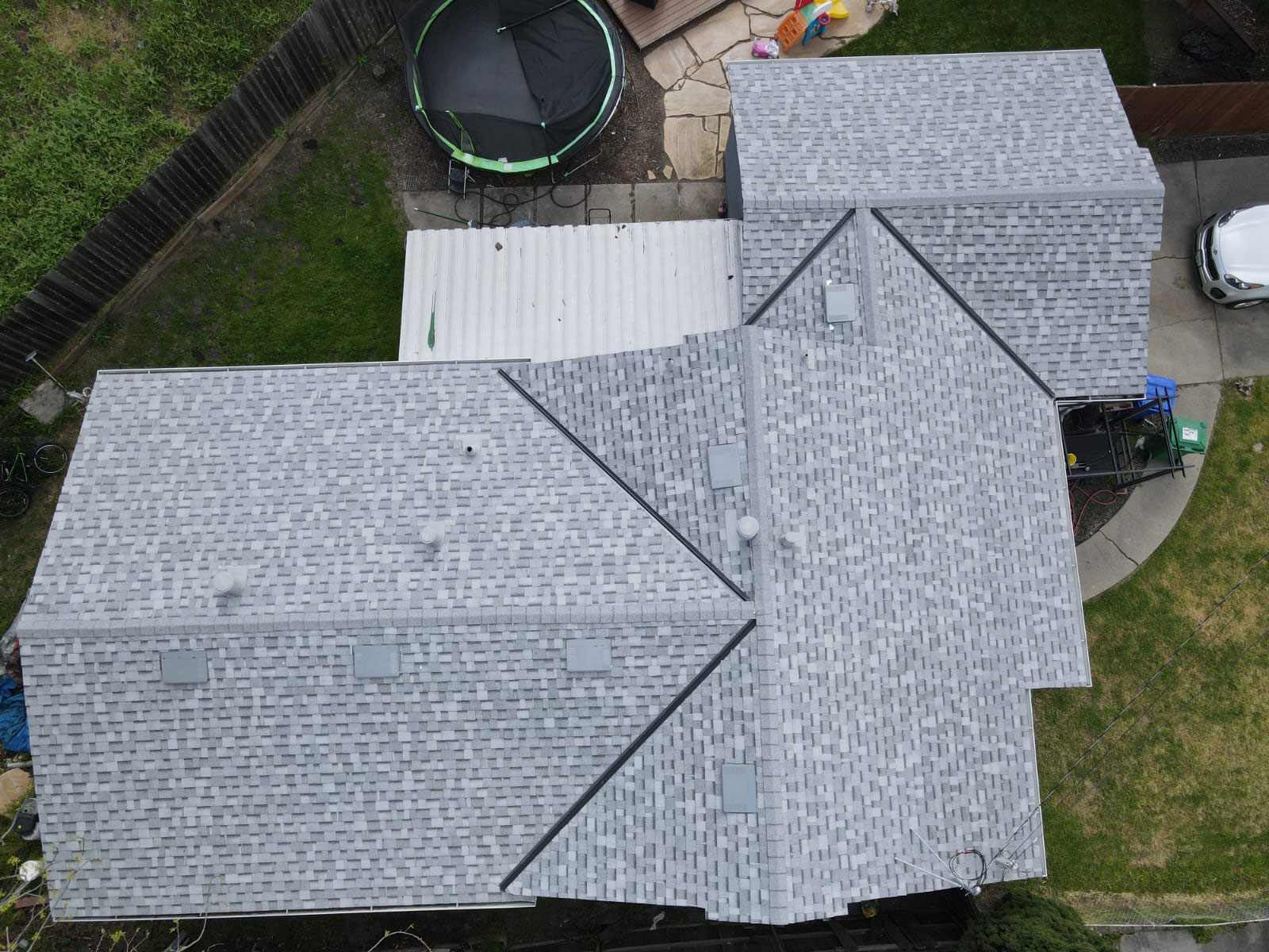 The image shows a residential house with a gray shingle roof, featuring a prominent triangular section at the front, situated on a street corner with a clear sky above.