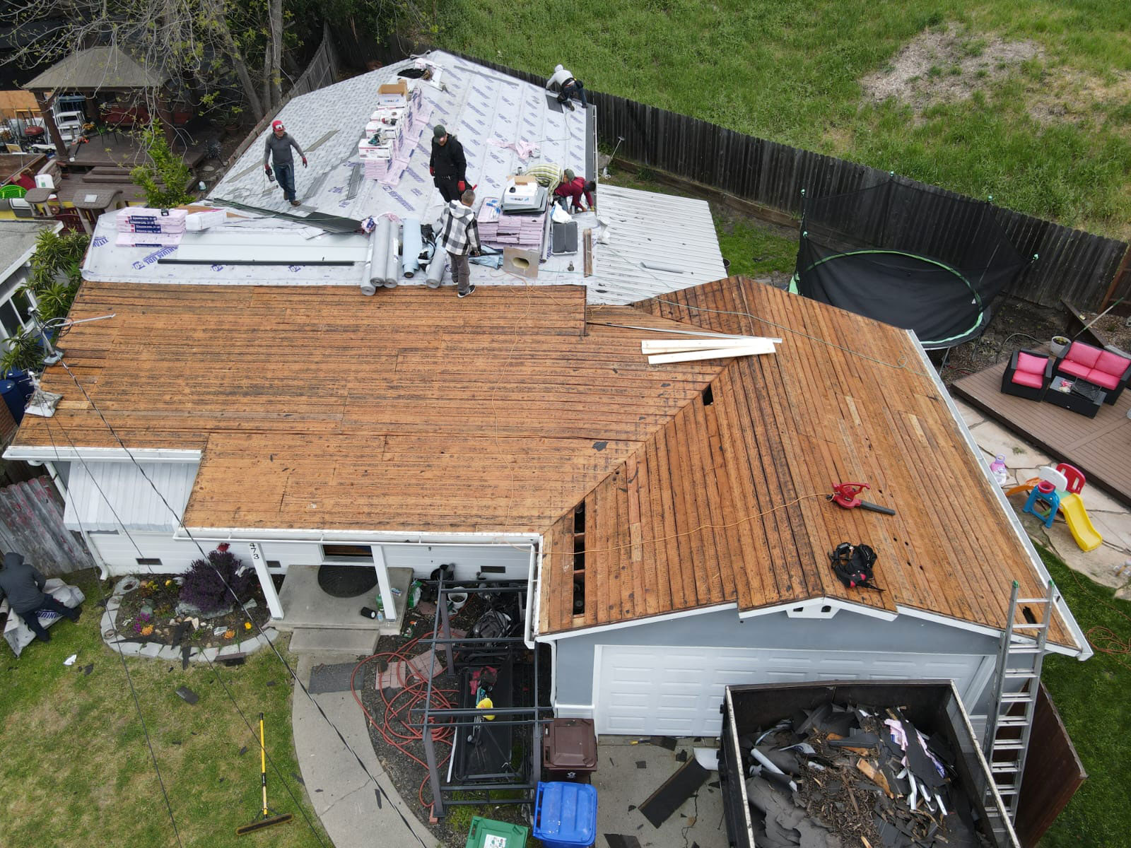 The image shows a house undergoing roof renovation with visible wooden planks on the upper level and construction materials scattered around the property.