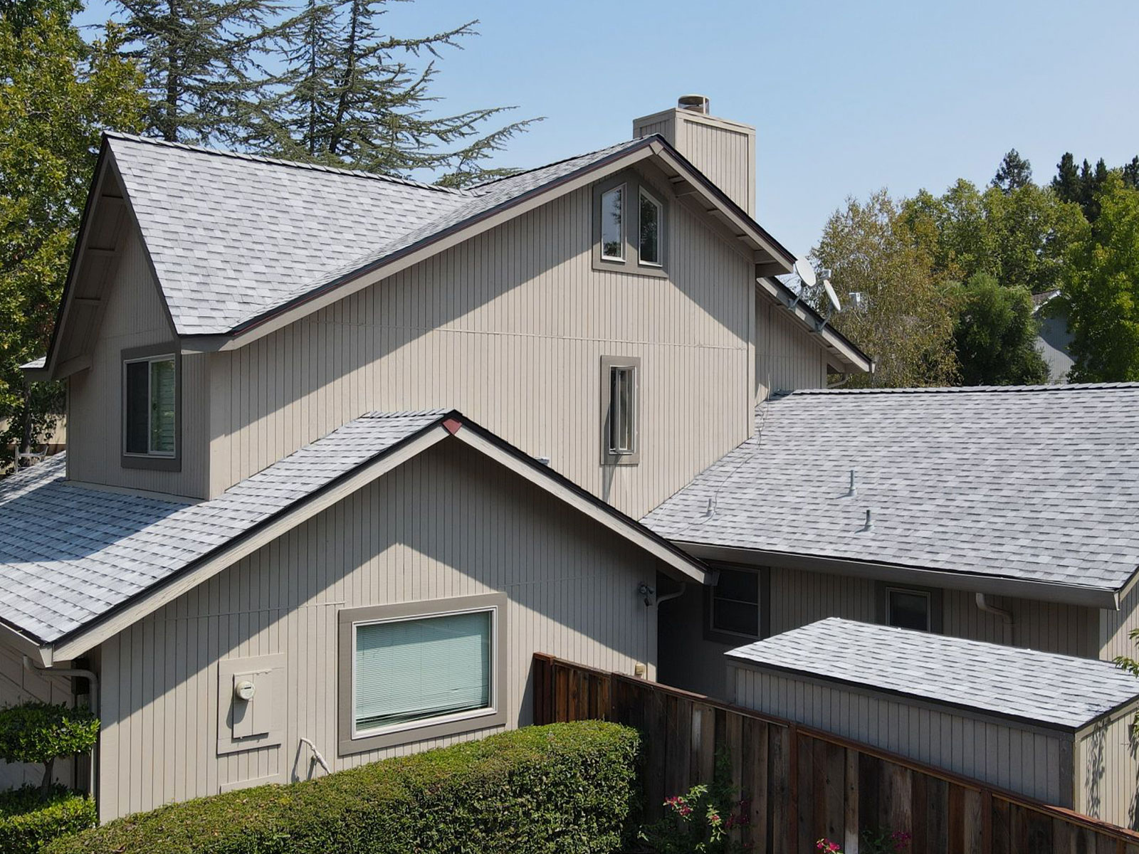 The image shows a two-story residential house with a gray roof and beige siding, featuring a garage and a front porch, set against a clear sky.