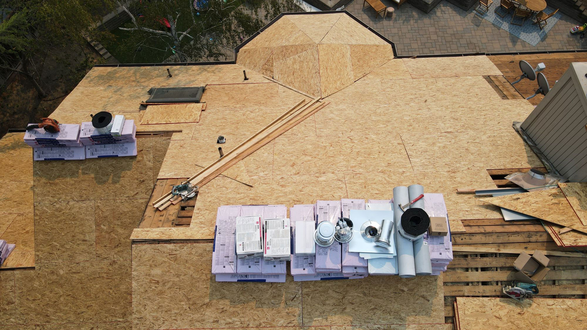 The image shows an aerial view of a partially constructed building with visible wooden framing, insulation, and construction materials on the ground around it.