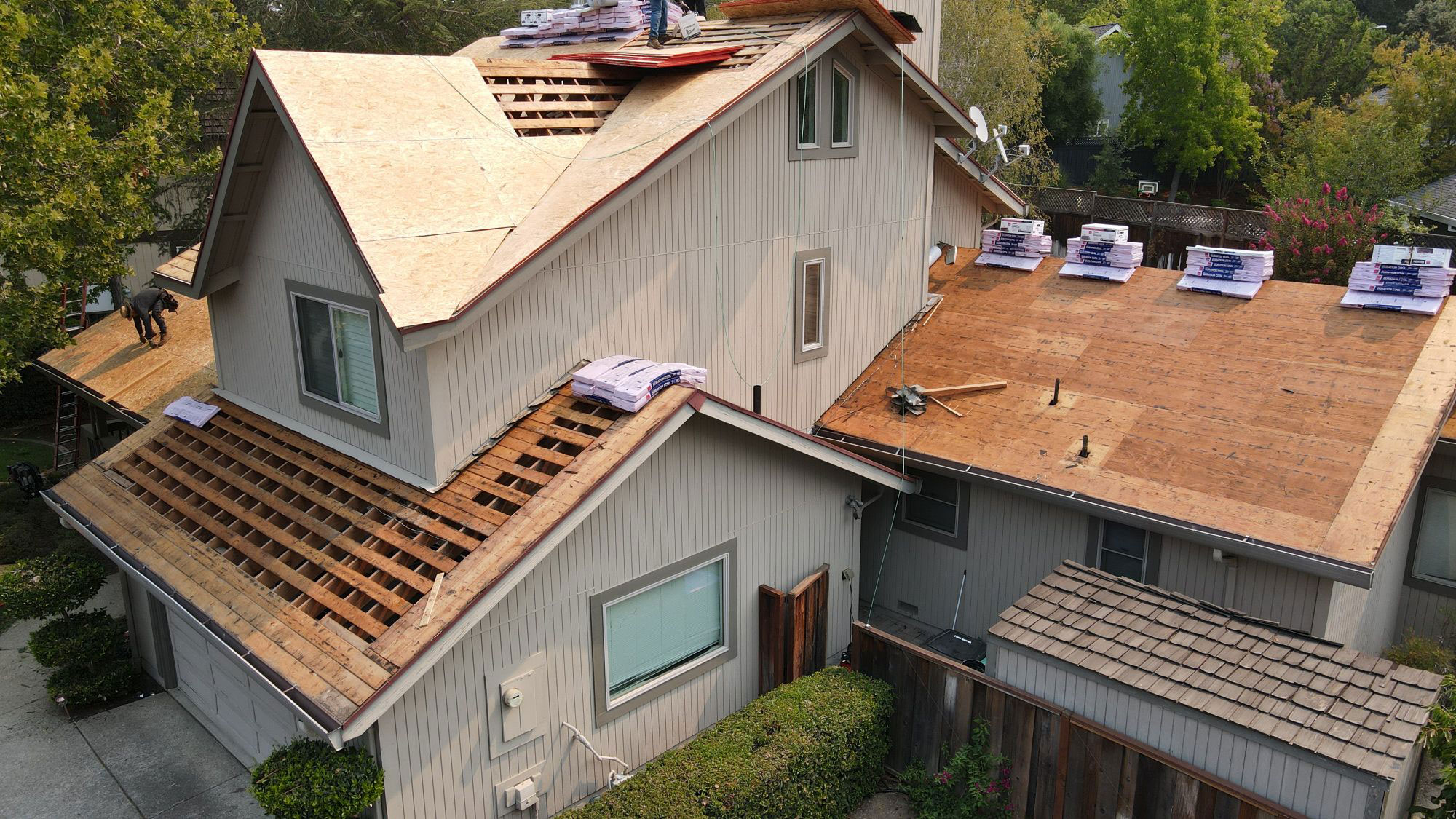 This image shows a residential house with a visible roof repair underway, characterized by exposed wooden decking and a variety of construction materials and equipment on the roof.