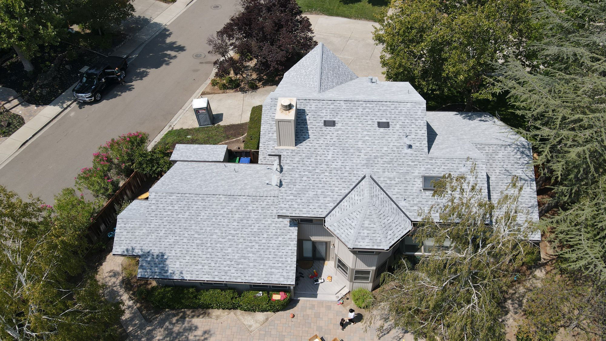 The image shows a two-story residential house with a shingled roof, featuring multiple gables and dormers, surrounded by a well-manicured lawn and landscaping.