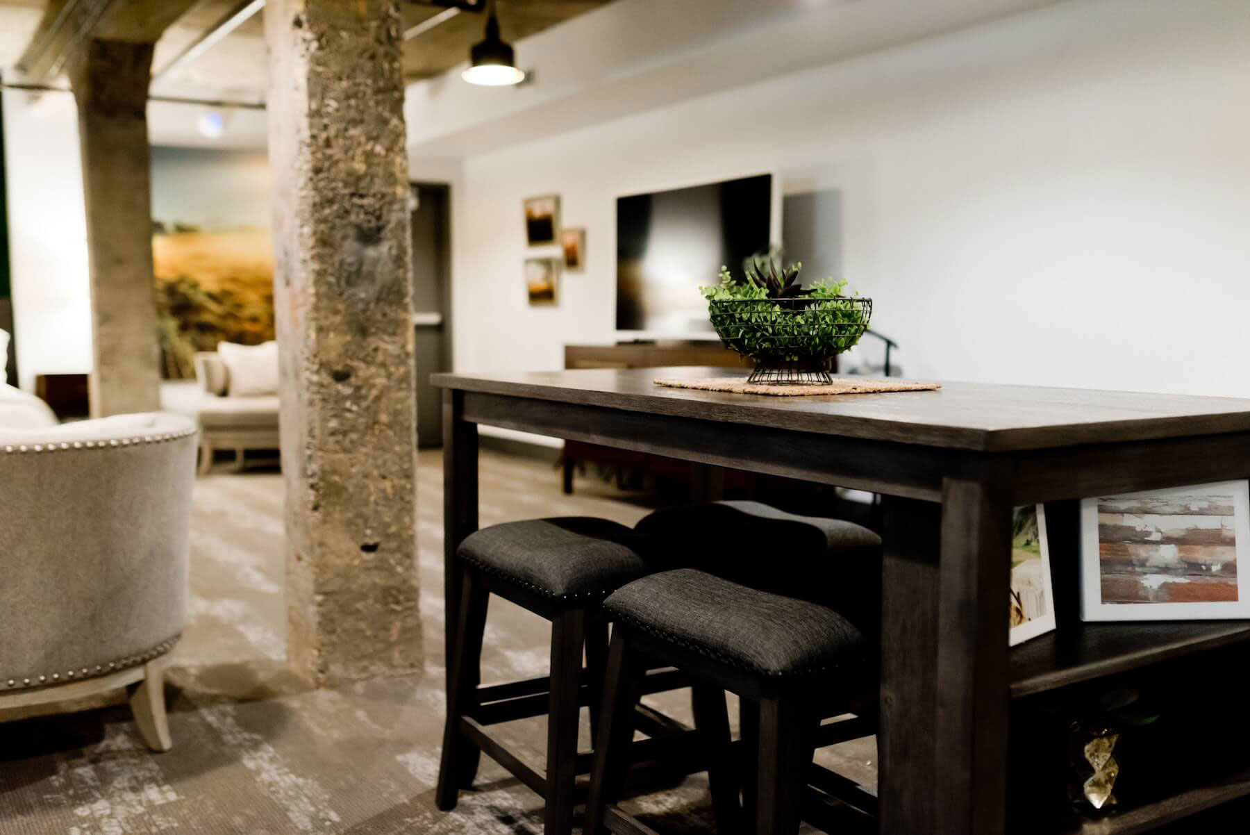 The image depicts a modern living room interior with a large wooden dining table at its center, surrounded by bar stools and high-top chairs, set against a backdrop of exposed brick walls and contemporary furnishings.