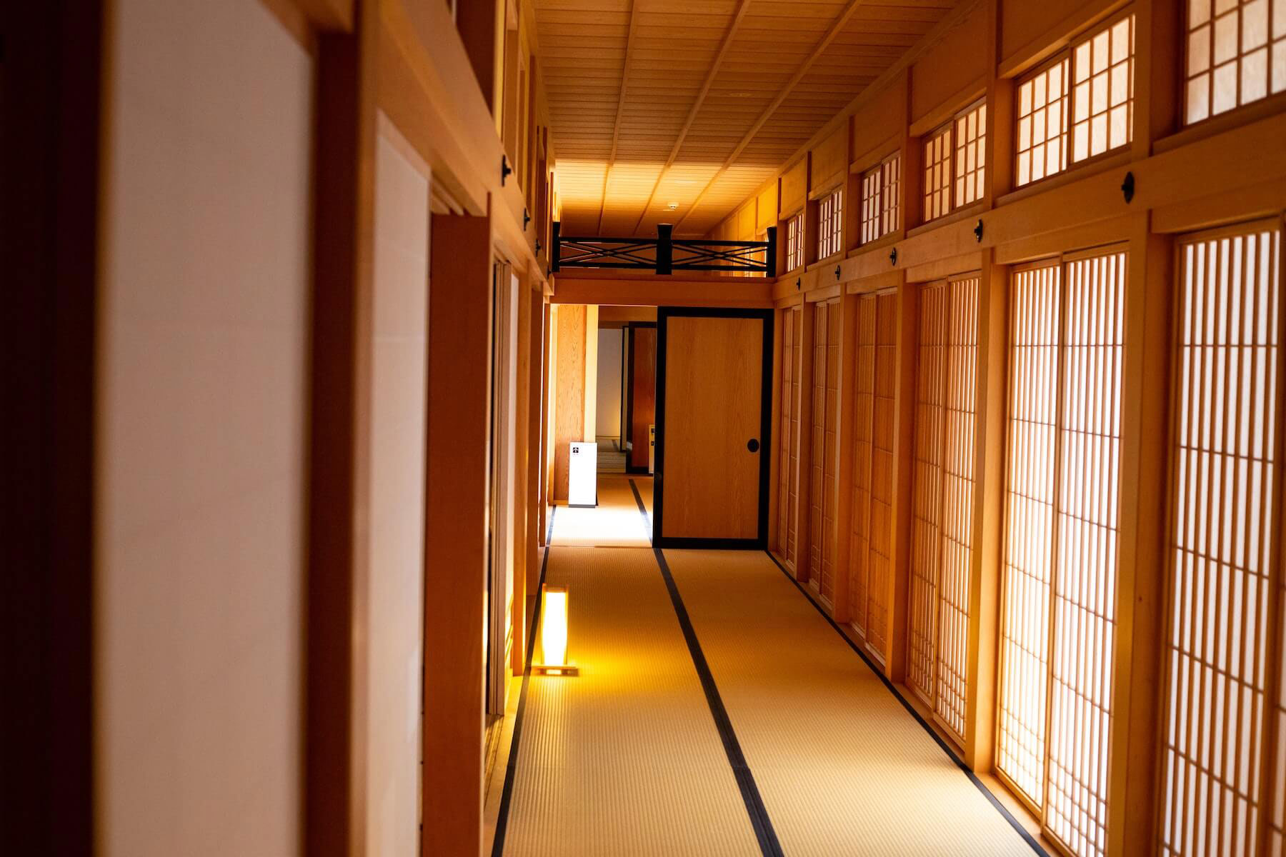 The image shows an interior hallway within a traditional Japanese ryokan with tatami flooring, sliding doors, and minimalist decor.